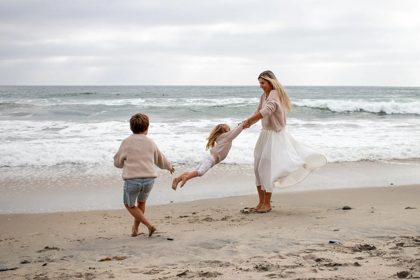 A woman and two children playing on the beach near the ocean, with the woman holding the girl in the air and another child walking nearby.
