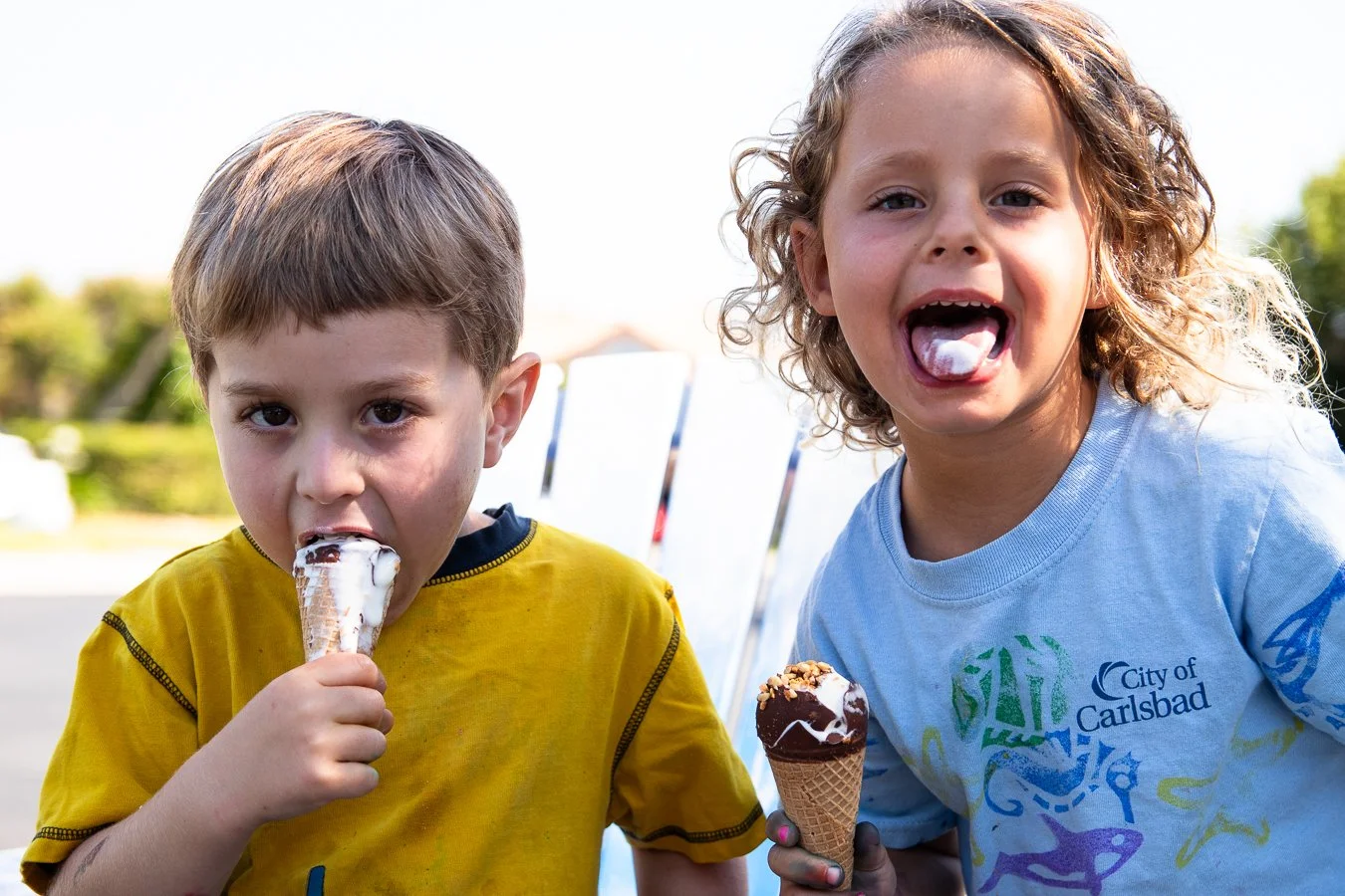 Two children, a boy in a yellow shirt and a girl in a light blue shirt, are outside enjoying ice cream cones on a sunny day.