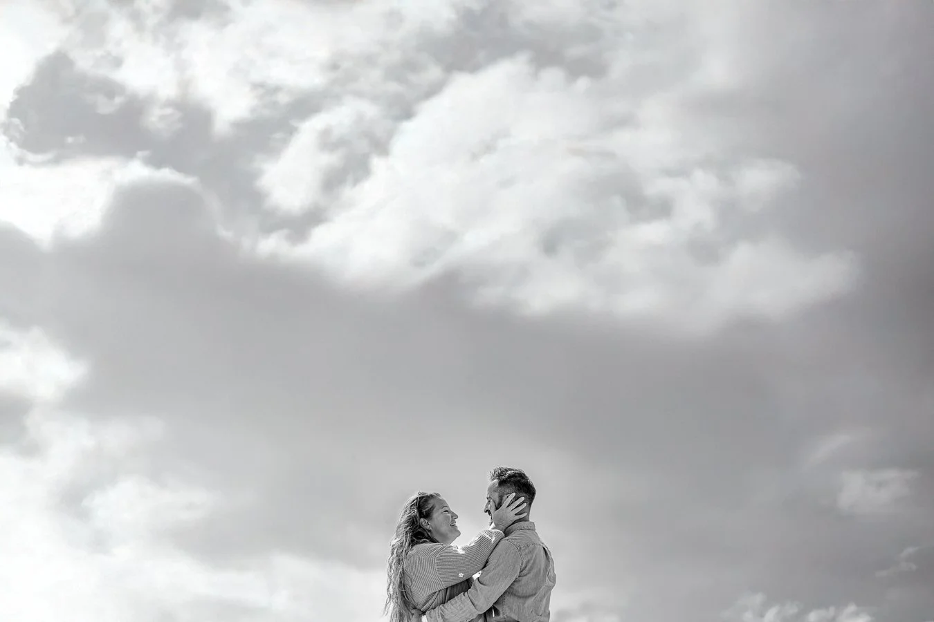 A black and white photo of a couple embracing outdoors under a cloudy sky.