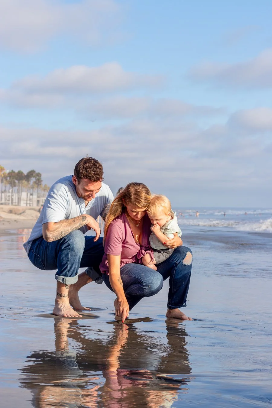 A family of three, with a man, woman, and young boy, exploring the shoreline at the beach during daytime. They are squatting at the water's edge, looking at the reflection and small creatures in the shallow water, with a cloudy sky and distant beachg