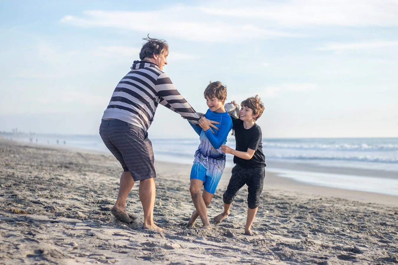 An adult man playing on the beach with two boys, all laughing and dressed in casual clothes, near the shoreline with the ocean and sky in the background.