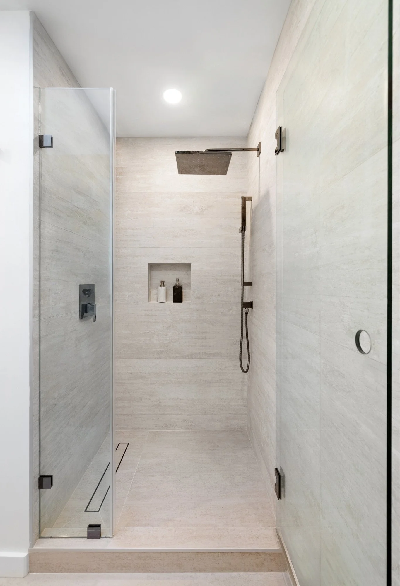 Modern walk-in shower with beige tiles, a large square shower head, a hand-held shower, a built-in niche with bottles, and a glass door.