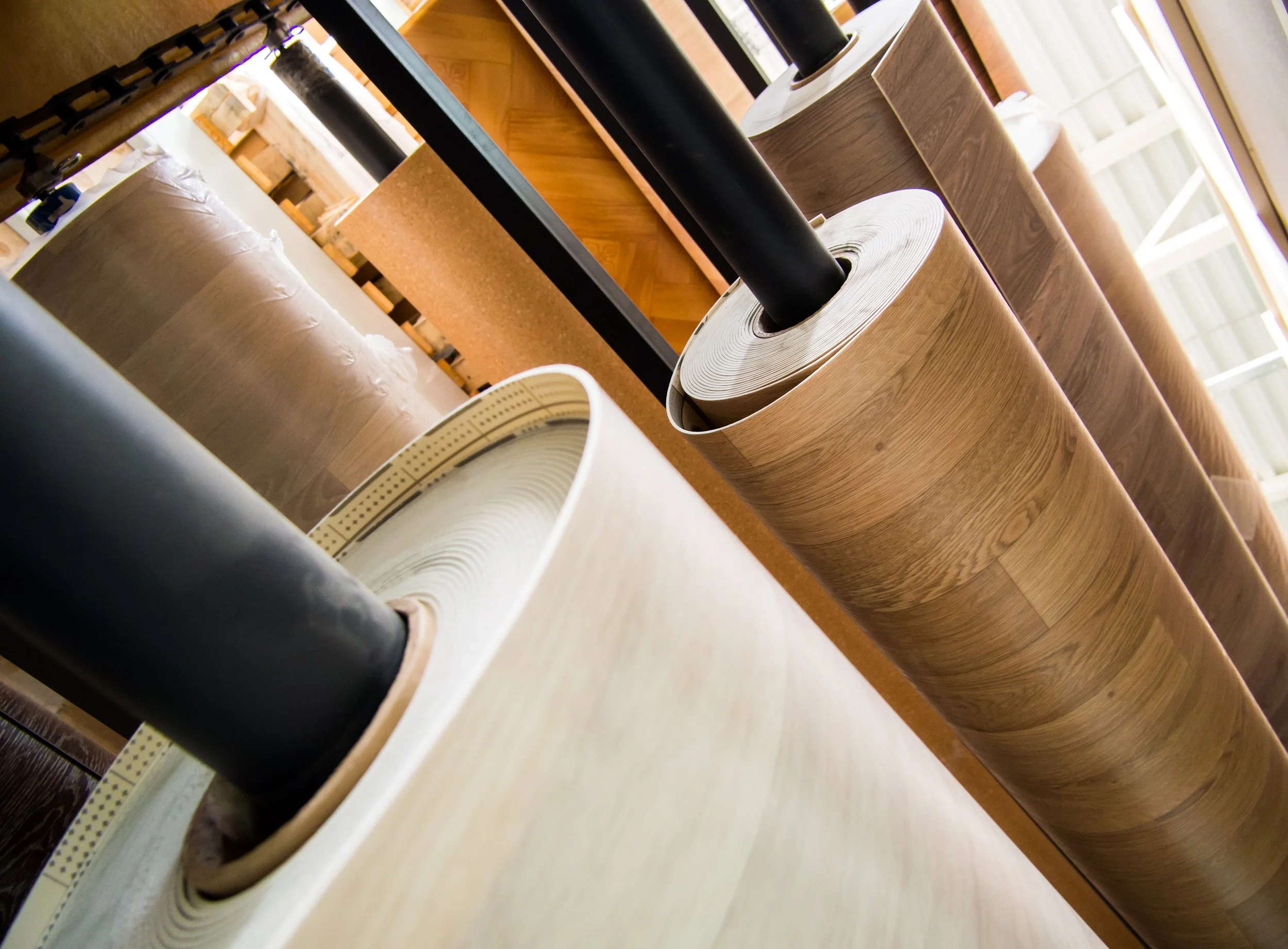 Close-up of several rolled-up carpets or rugs placed on their sides in a room with windows and wooden flooring.