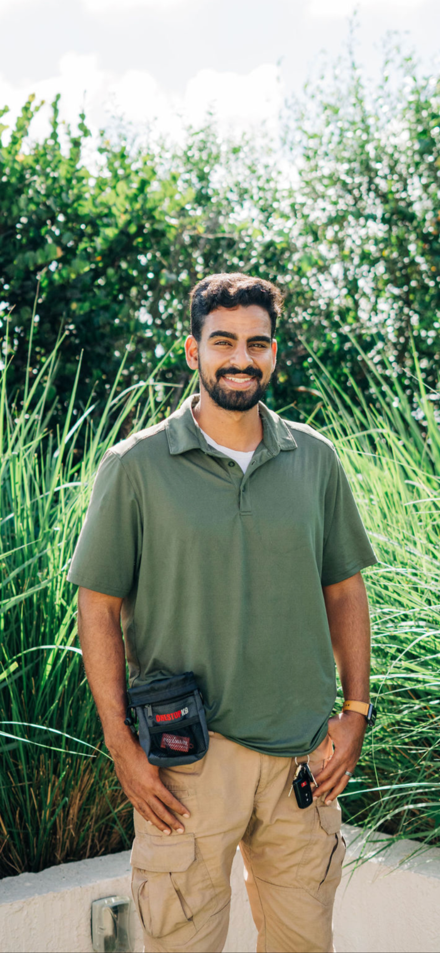 A man with dark hair and a beard standing outdoors in front of tall green grass and leafy bushes, smiling at the camera, wearing a green polo shirt, beige cargo pants, a wristwatch, and a small black pouch at his waist.