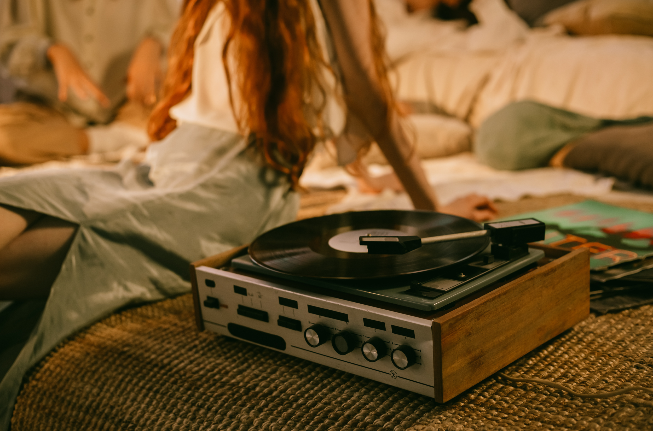 A person with long red hair lying on a bed in a cozy, softly lit room with a turntable and vinyl records nearby.