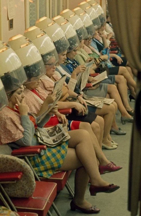 A row of women sitting in chairs with hair dryers on their heads, reading magazines in a beauty salon or spa.