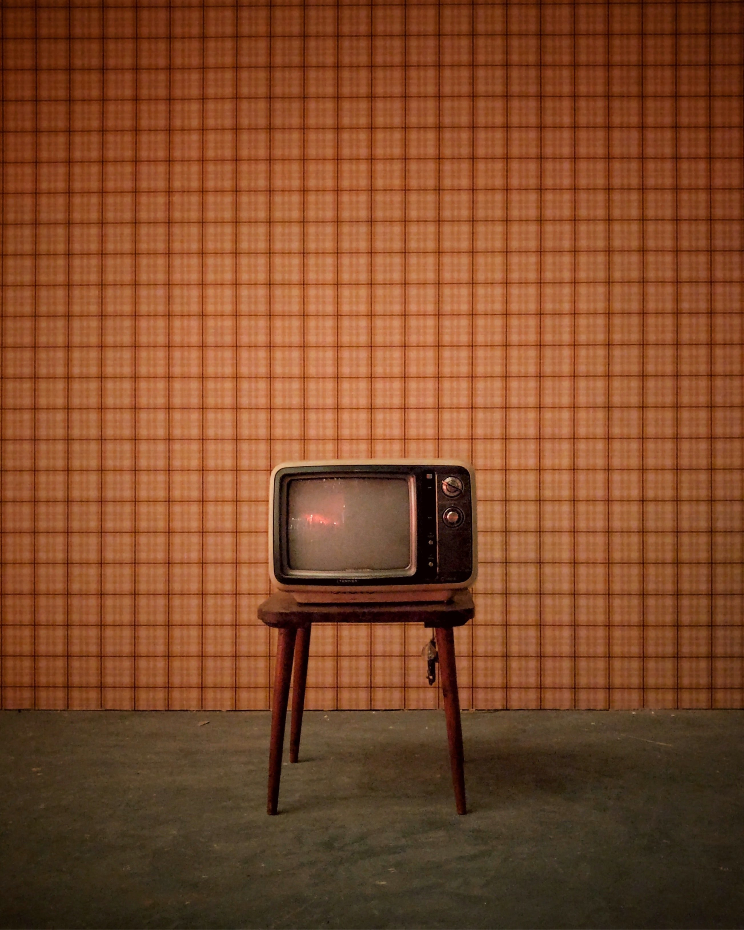 Old-fashioned television set on a wooden stand against a checkered wallpapered wall in a dimly lit room.
