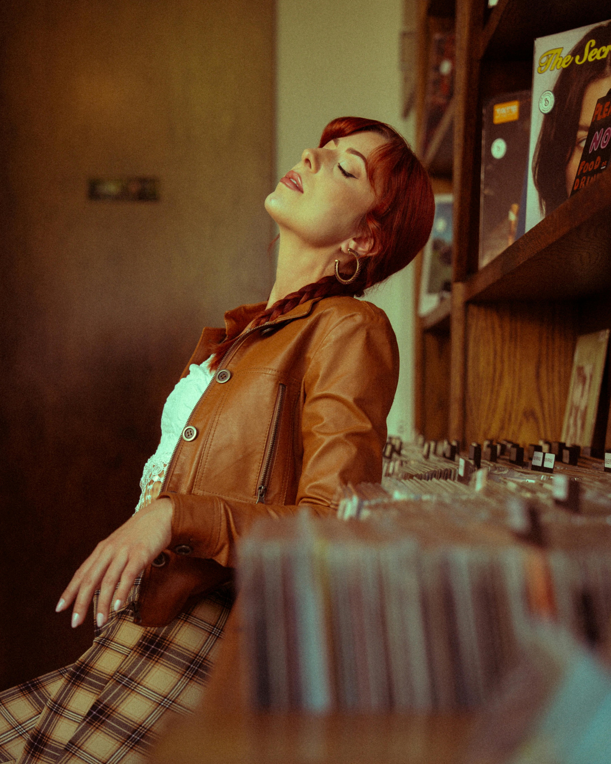 A woman with red hair and hoop earrings, wearing a brown leather jacket and plaid pants, leaning back with eyes closed in a record store, with vinyl records and posters visible in the background.