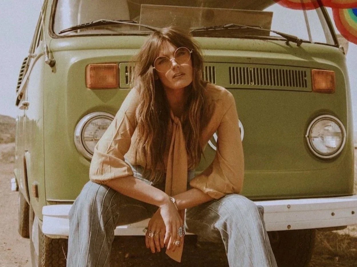 A woman with long hair and round glasses sitting on the front bumper of a vintage green van, with desert landscape in the background.