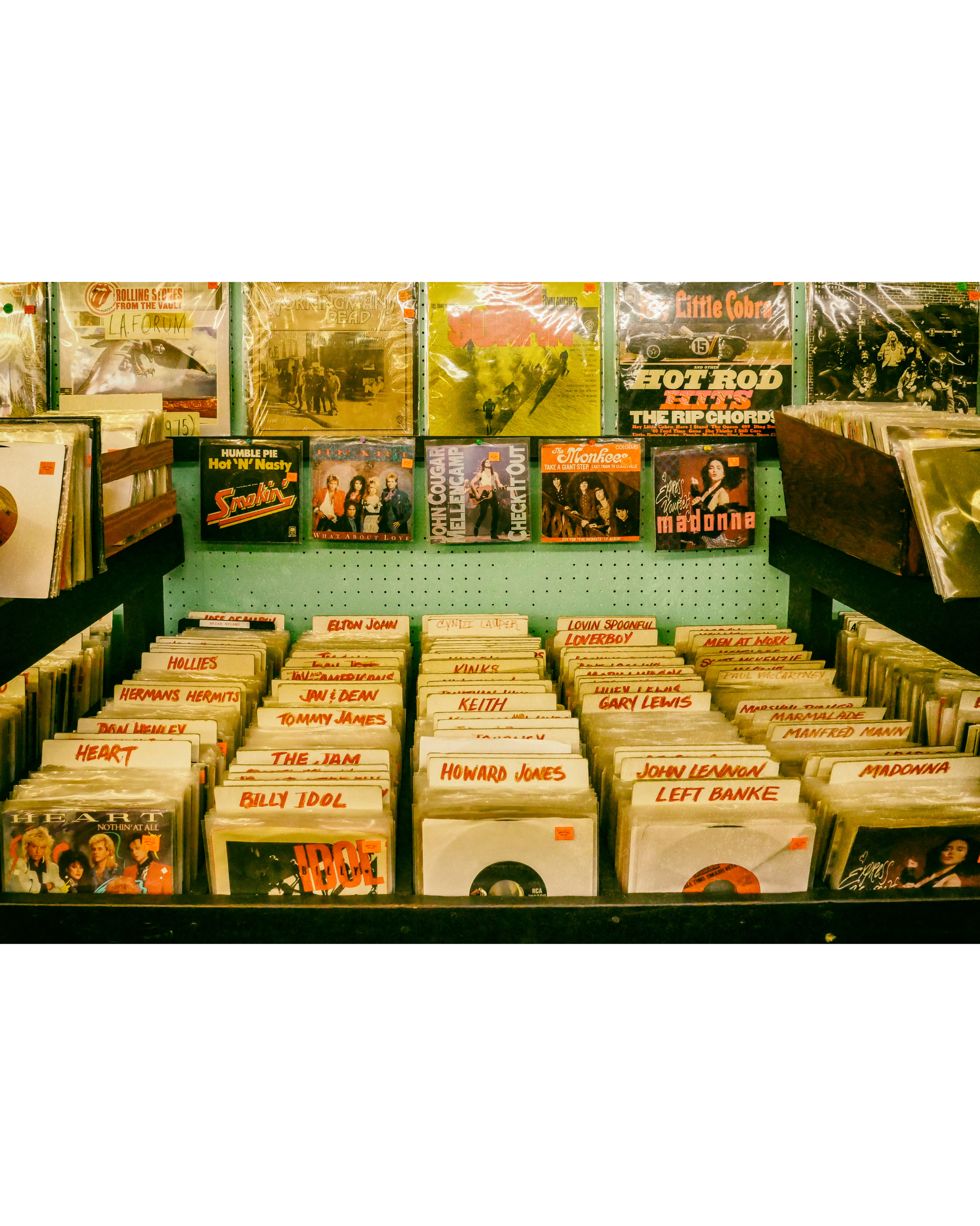 A vintage vinyl record store shelf displaying numerous records organized alphabetically, with the top row featuring classic rock albums and the lower row containing records by various artists, including Madonna and John Lennon.