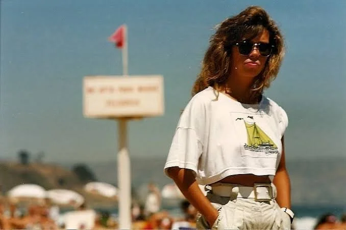 Woman with wavy hair wearing sunglasses, a white cropped t-shirt with a sailboat graphic, and beige high-waisted shorts, standing on a beach with umbrellas and a sign in the background.