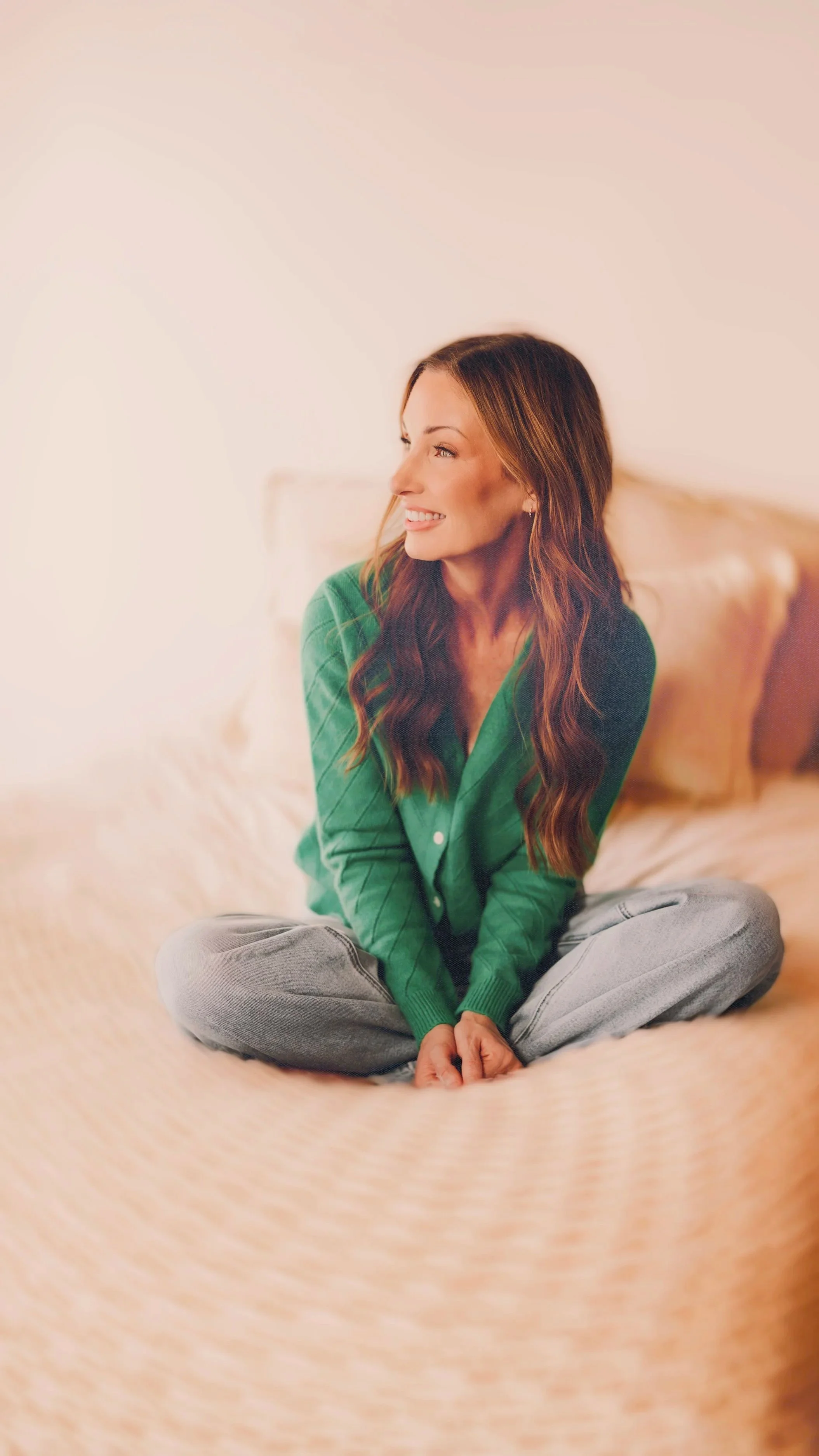 A woman with long wavy brown hair sitting on a bed with a beige headboard, wearing a green cardigan and grey sweatpants, smiling and looking to the side.