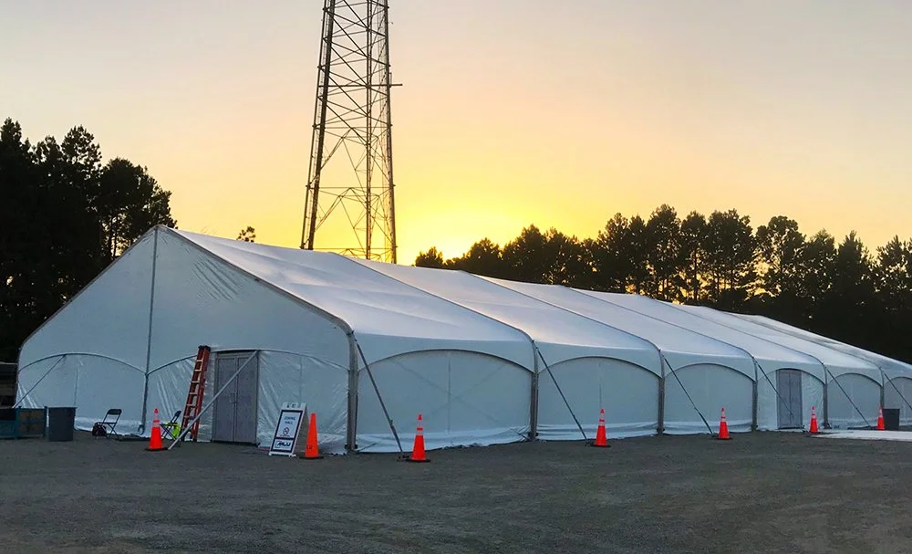Large white tent set up outdoors during sunset, with orange traffic cones in front and a tall metal tower behind. Trees are visible in the background.