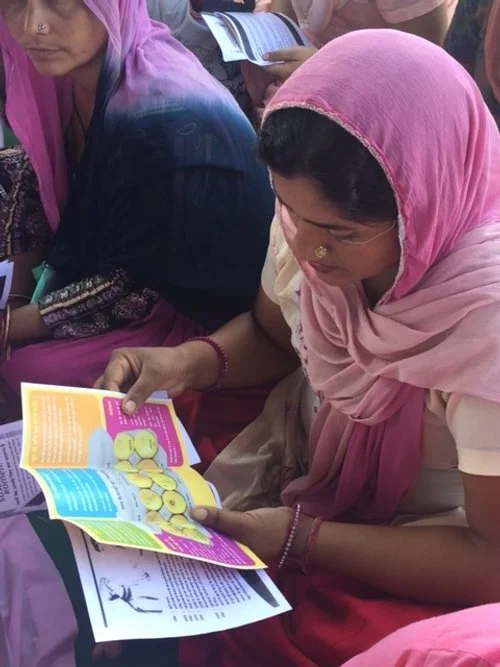 Woman wearing a pink headscarf reading a colorful brochure or pamphlet during a gathering or event, with other women nearby.