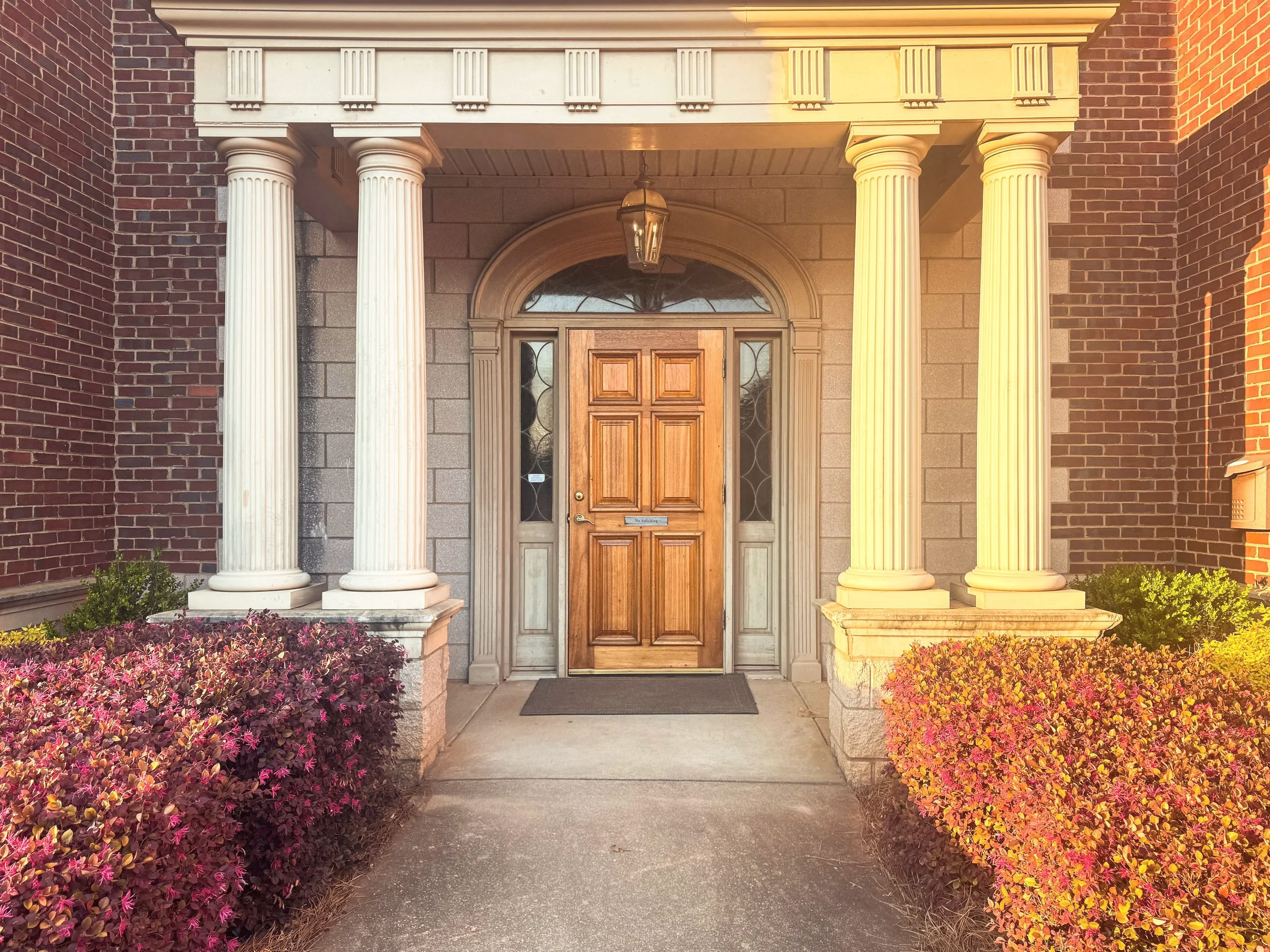 Front door of a house with four tall white columns, a wooden door, potted plants on either side, and bushes with purple and yellow leaves.