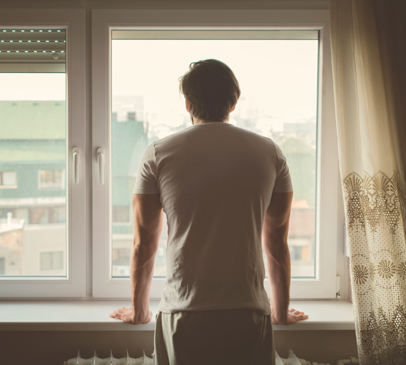 A man standing by a window, looking outdoors, with his back facing the camera. The window has a white frame and the room has light-colored curtains with a lace pattern.