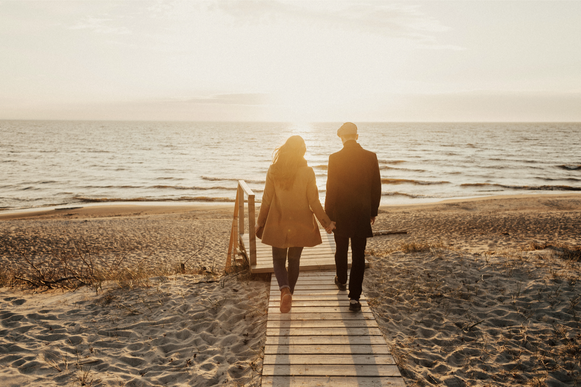 A man and woman walking through a wheat field with the man gently holding the woman from behind on a cloudy day.