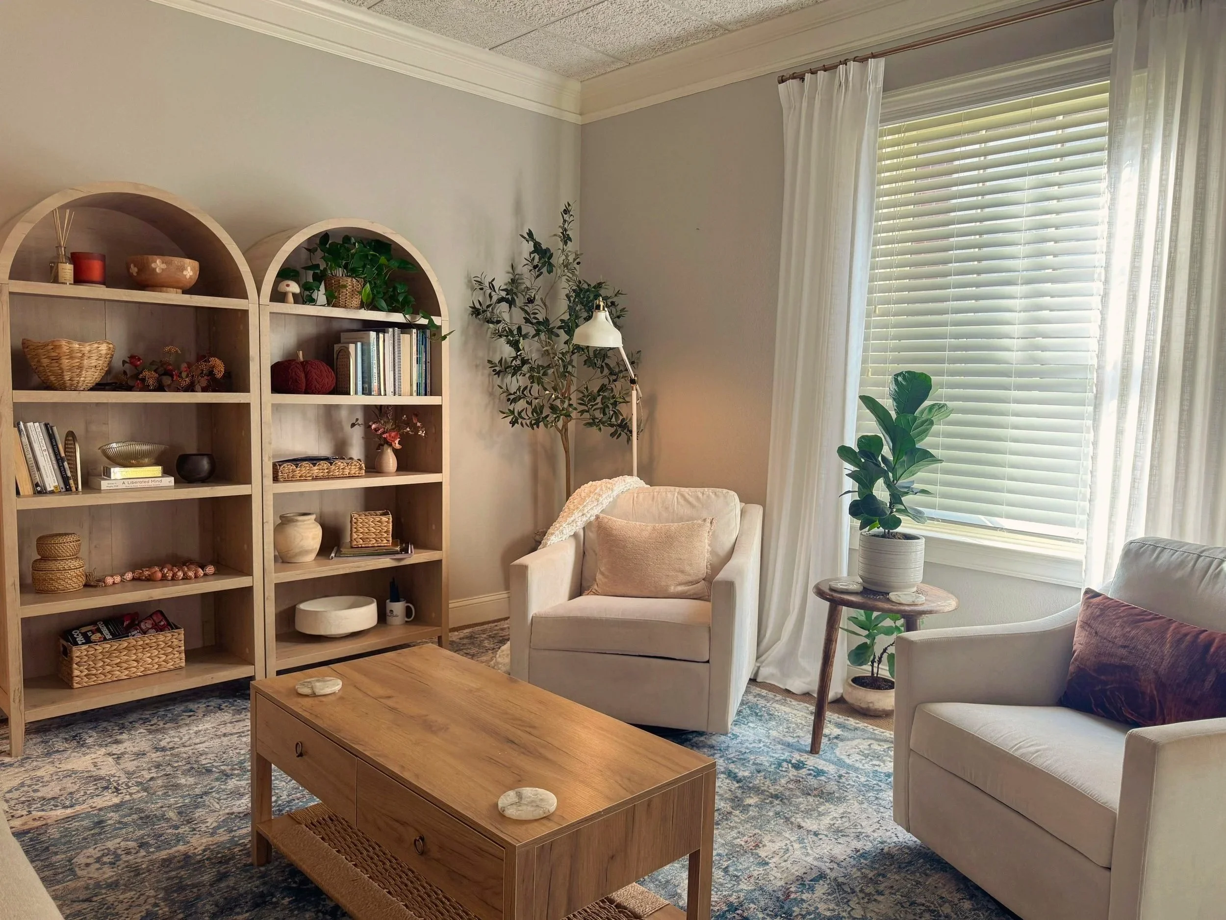 Office space with two white armchairs, a wooden coffee table, a tall bookshelf with decorative items, a potted plant, and a window with blinds and curtains.