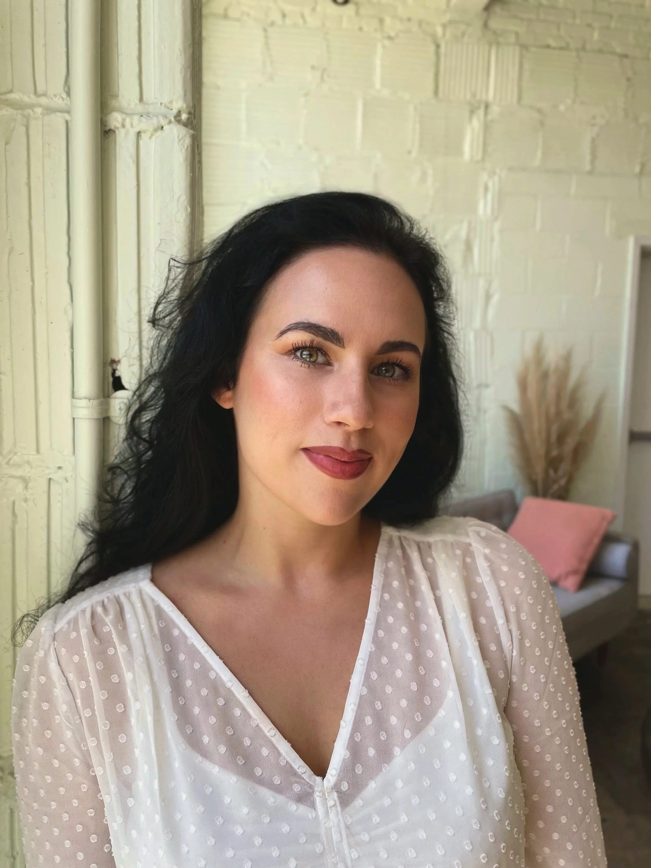 A woman with dark hair, light skin, and makeup, wearing a white polka-dotted blouse, standing indoors near a brick wall with a beige couch and pink pillow in the background.