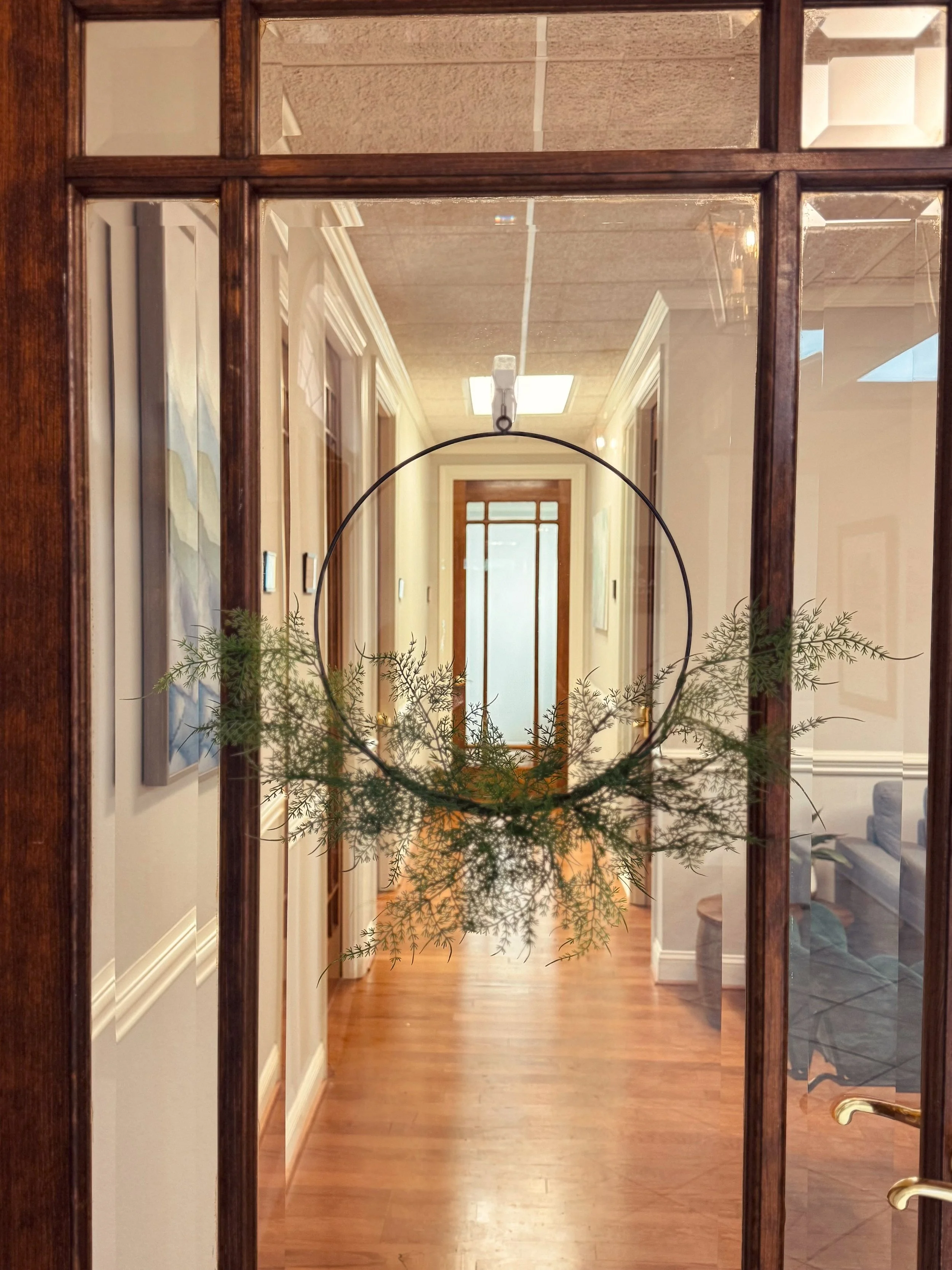 A hallway viewed through a glass door with a wreath hanging in the center, decorated with greenery. The hallway has wooden floors, white walls, several framed artworks, and doors along the corridor, leading to a door at the end with light coming through.