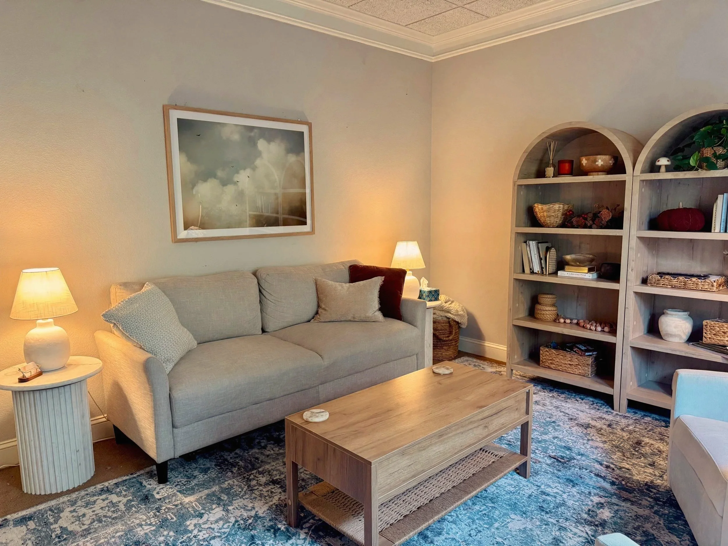 Room with beige sofa, wooden coffee table, patterned rug, two table lamps, framed wall art, and wooden bookshelves with decorative items.