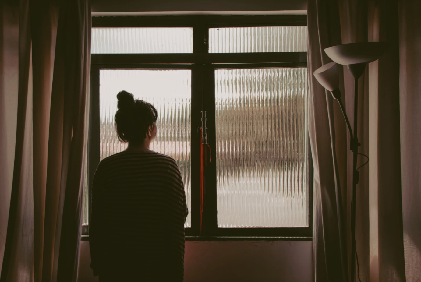 A woman with long, curly hair standing outdoors in sunlight, wearing a white crocheted shawl with fringe, arms outstretched, and eyes closed.