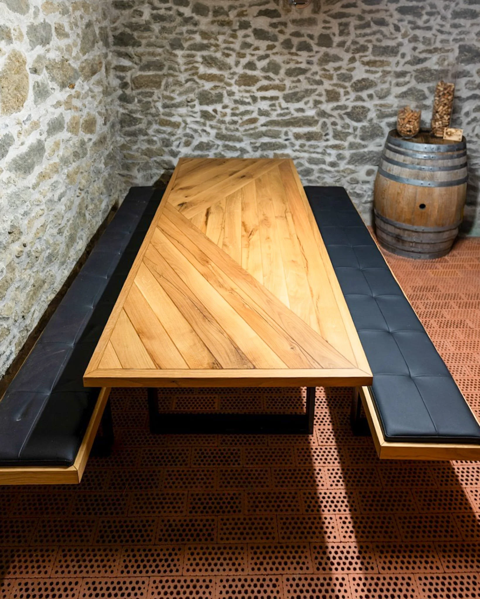 Table en bois avec bancs en cuir noir dans une pièce aux murs en pierre, sol en pavés en terre cuite, et un tonneau en bois avec des boules de bois dessus dans le coin