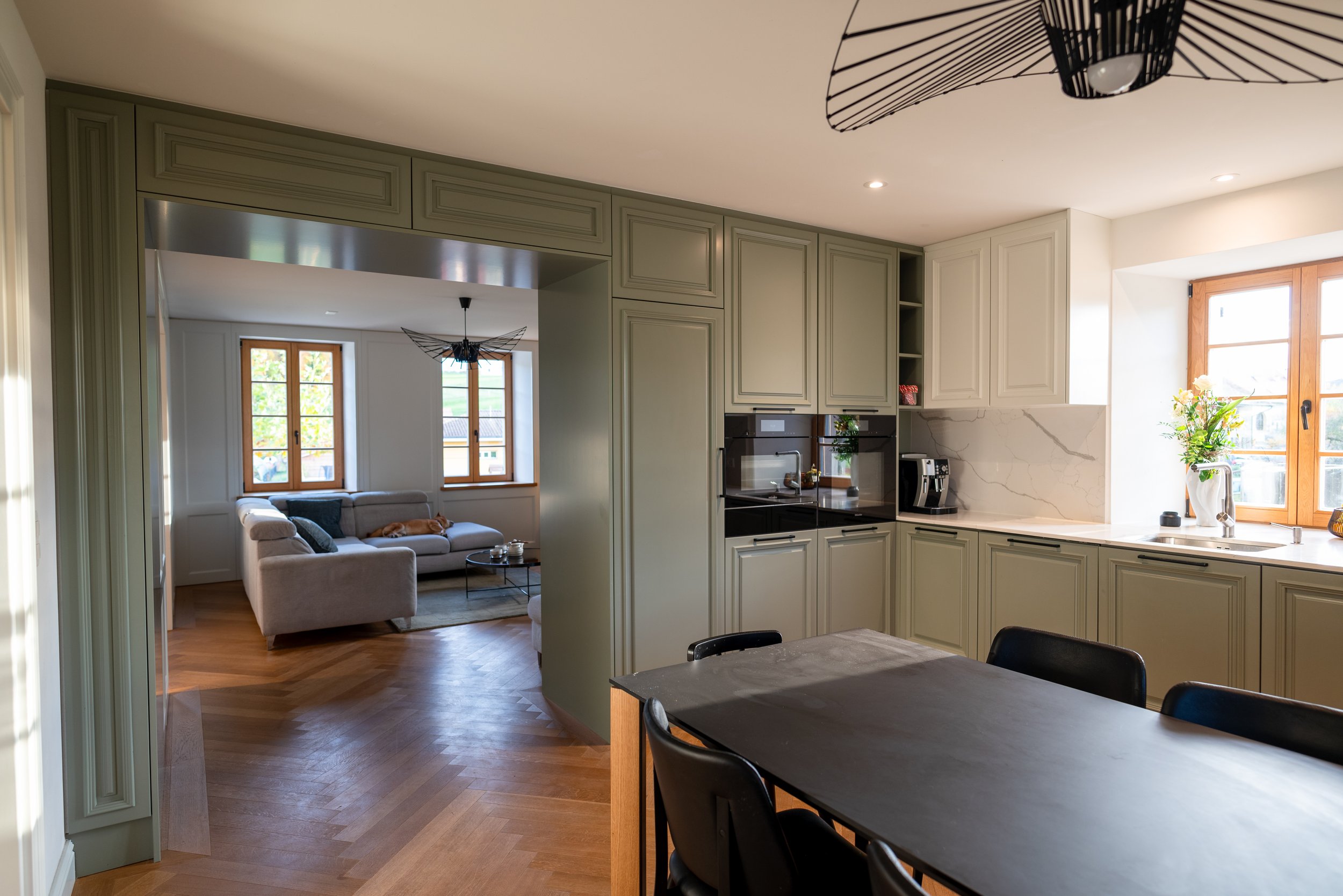 Cuisine ouverte avec armoires vert clair, plan de travail en marbre, fenêtre avec vue sur l'extérieur, table en bois noir avec sièges en cuir noir, pièce attenante avec canapé gris clair, tables basses, et fenêtres en bois clair laissant entrer la lumière naturelle.