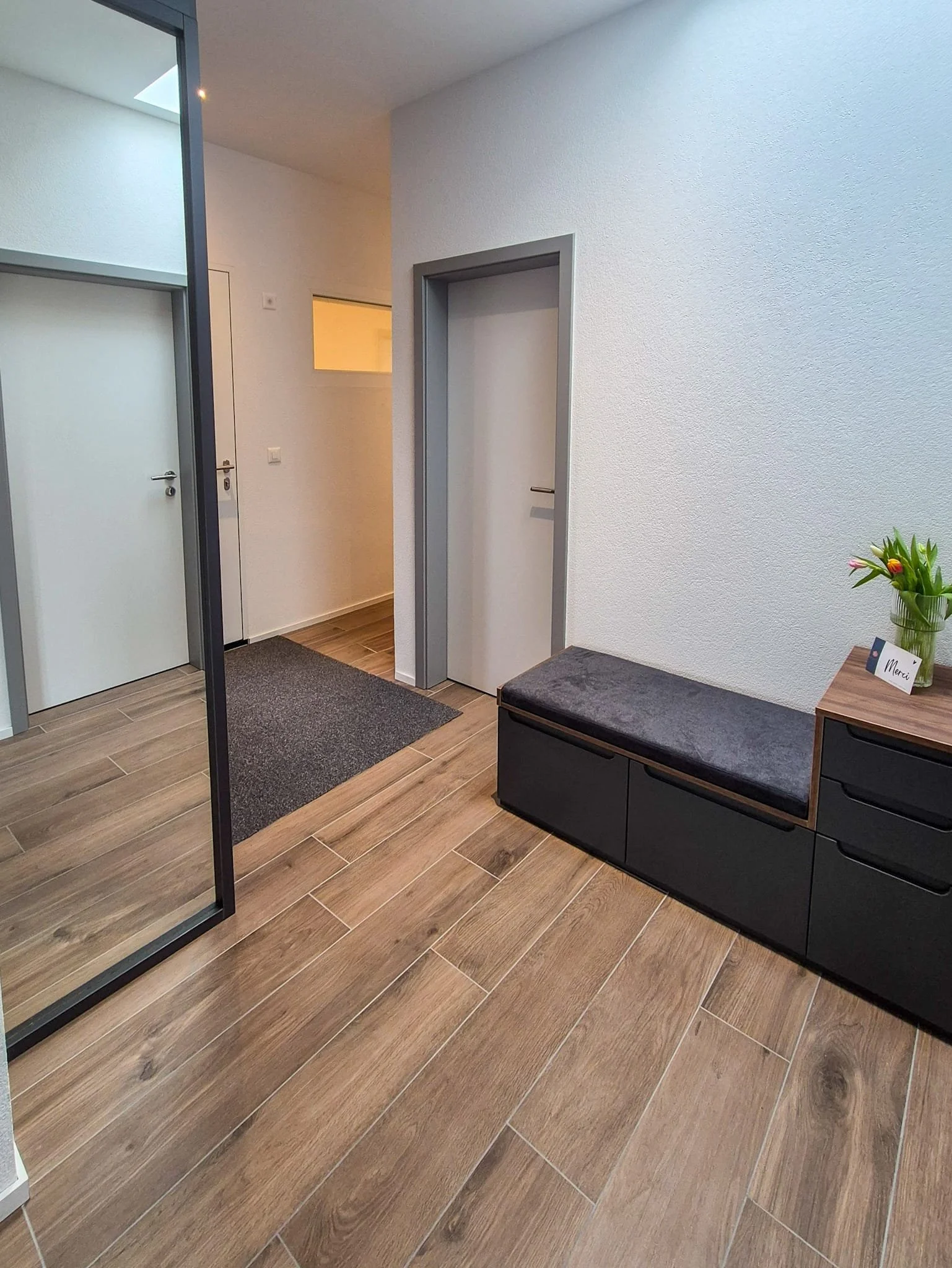 Intérieur moderne avec plancher en bois, porte d'entrée, banc noir avec espace de rangement, plante verte, et petites notes de bienvenue.