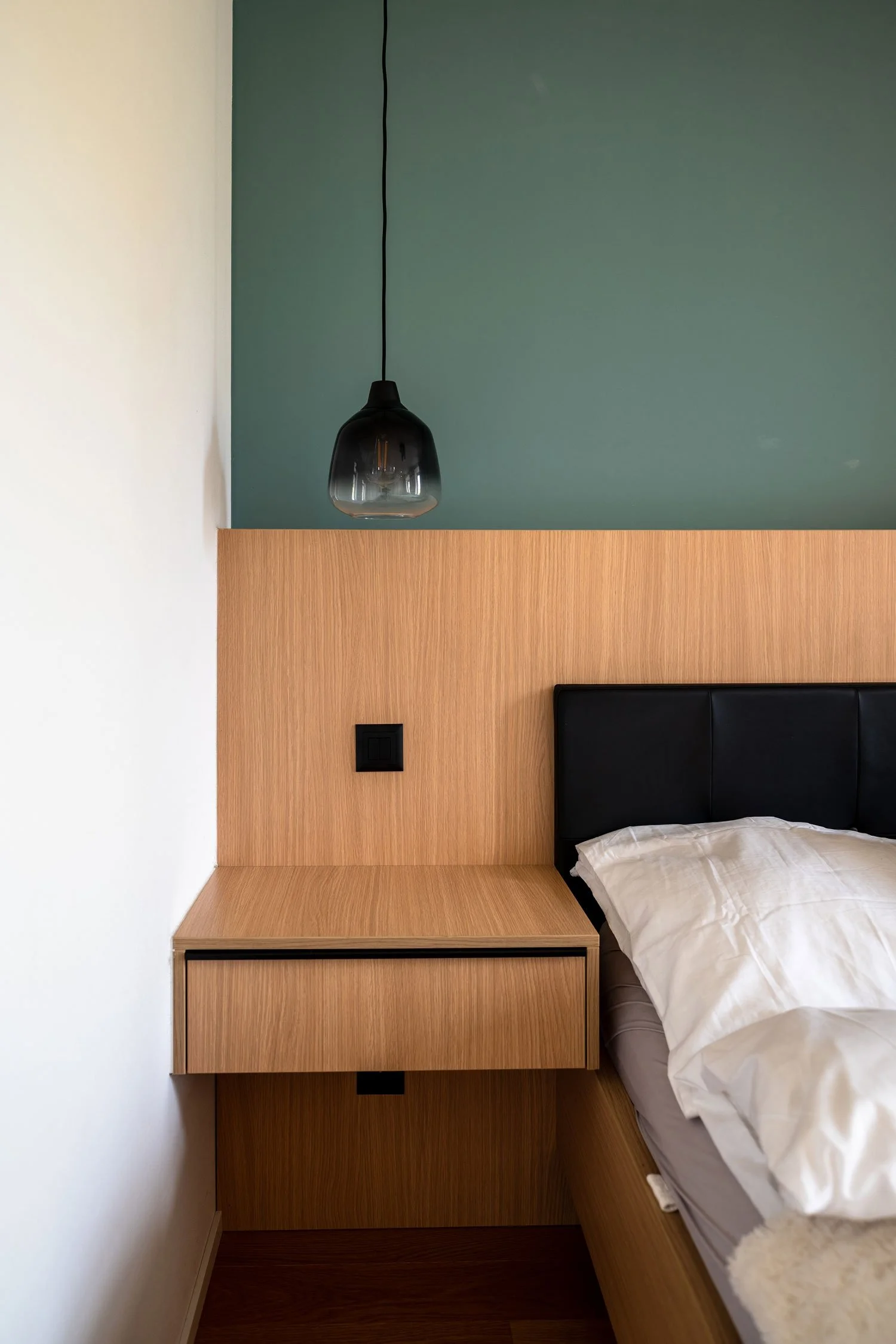 Chambre avec lit, chevet en bois, panneau en bois derrière le lit, peinture de mur vert foncé et blanc, lampe suspendue noire