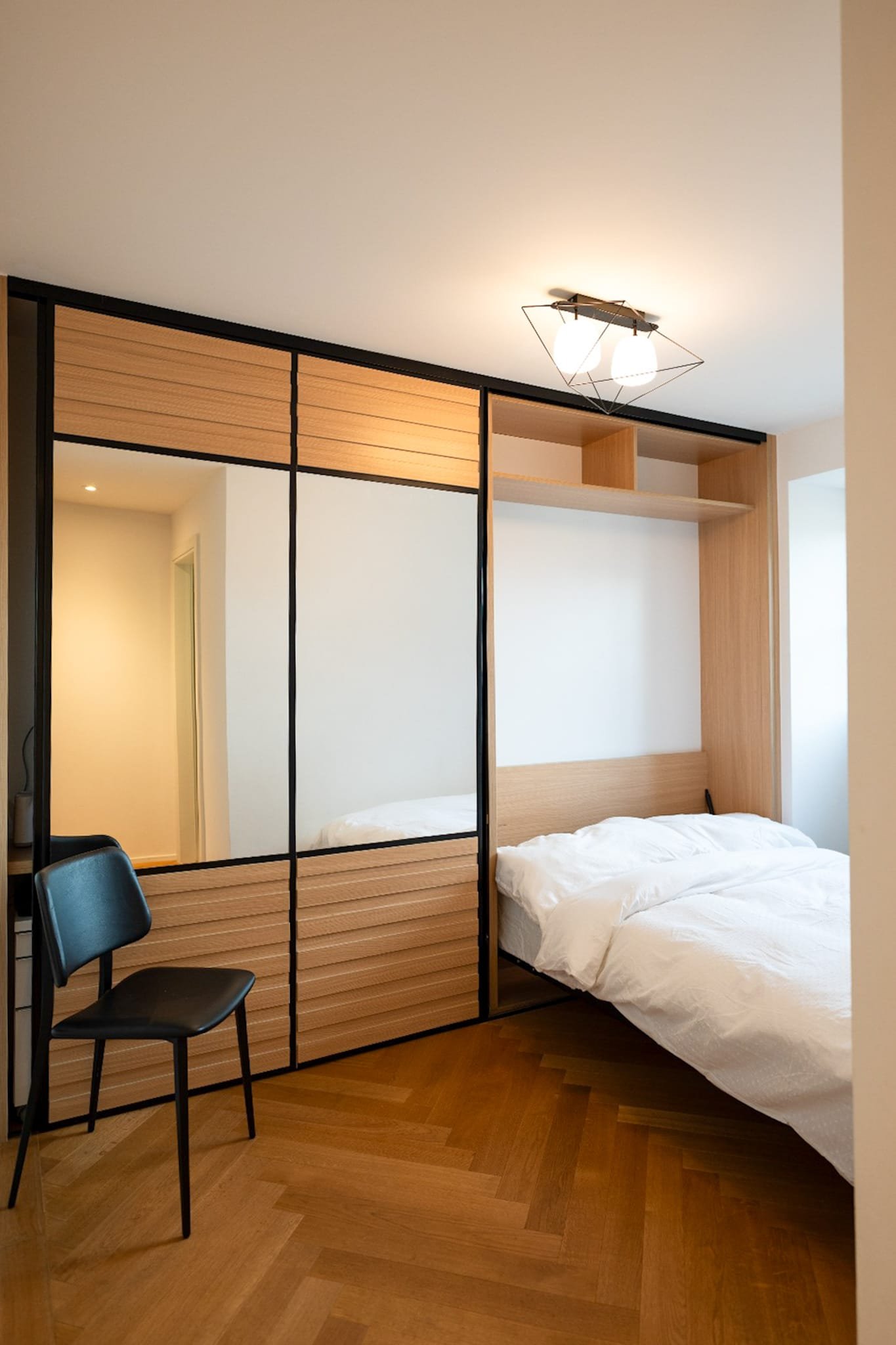 Chambre moderne avec lit blanc, grande armoire avec miroir et portes en bois, sol en bois, lampe suspendue à deux lumières.