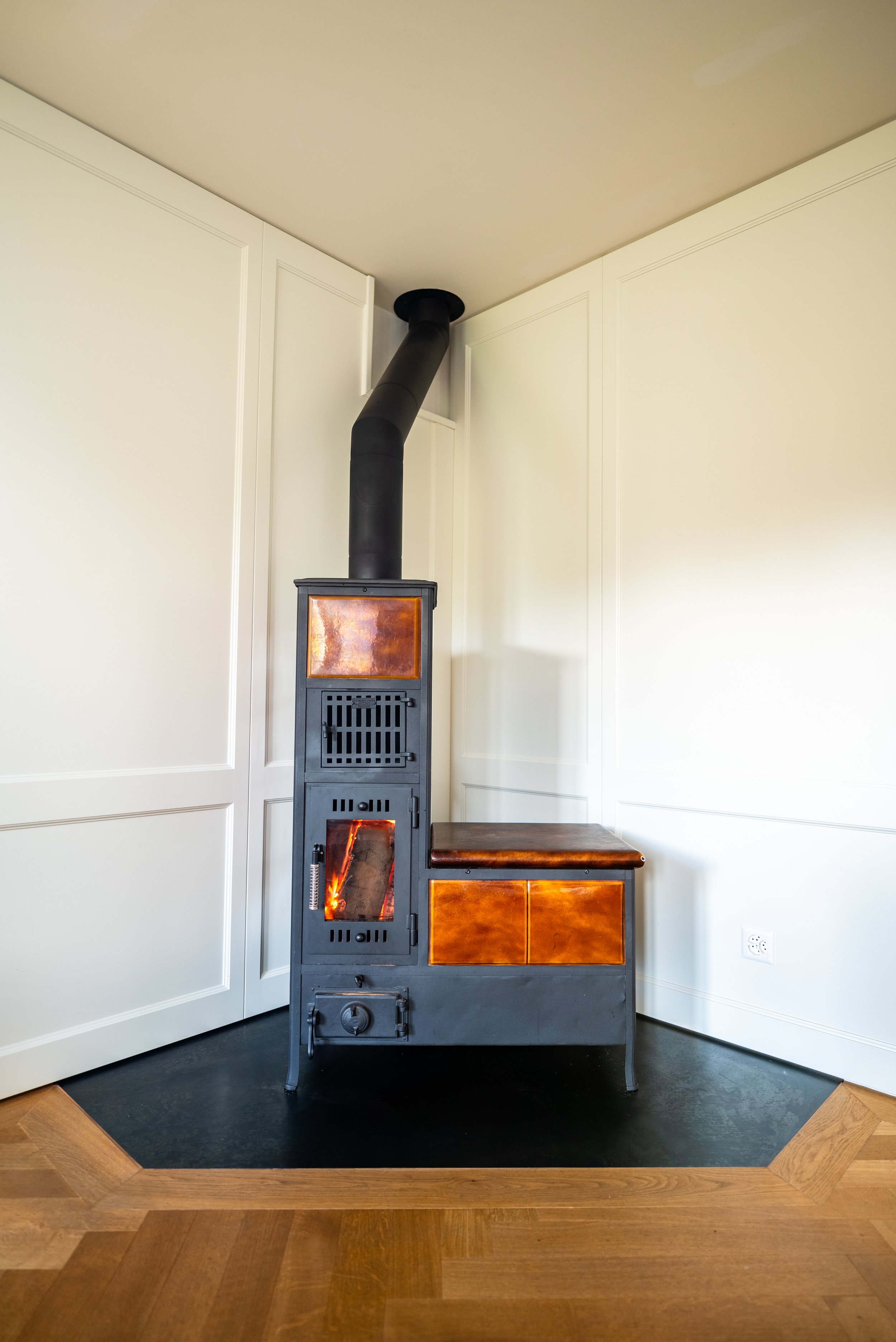 Poêle à bois noir avec porte vitrée en verre, situé dans un coin de pièce avec sol en bois et murs blancs.