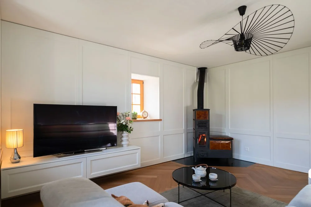 Salon avec une télévision noire, une cheminée, une table basse ronde, une lampe, un canapé clair, et une fenêtre avec une plante et une horloge. Mur blanc avec panneau décoratif, sol en parquet.