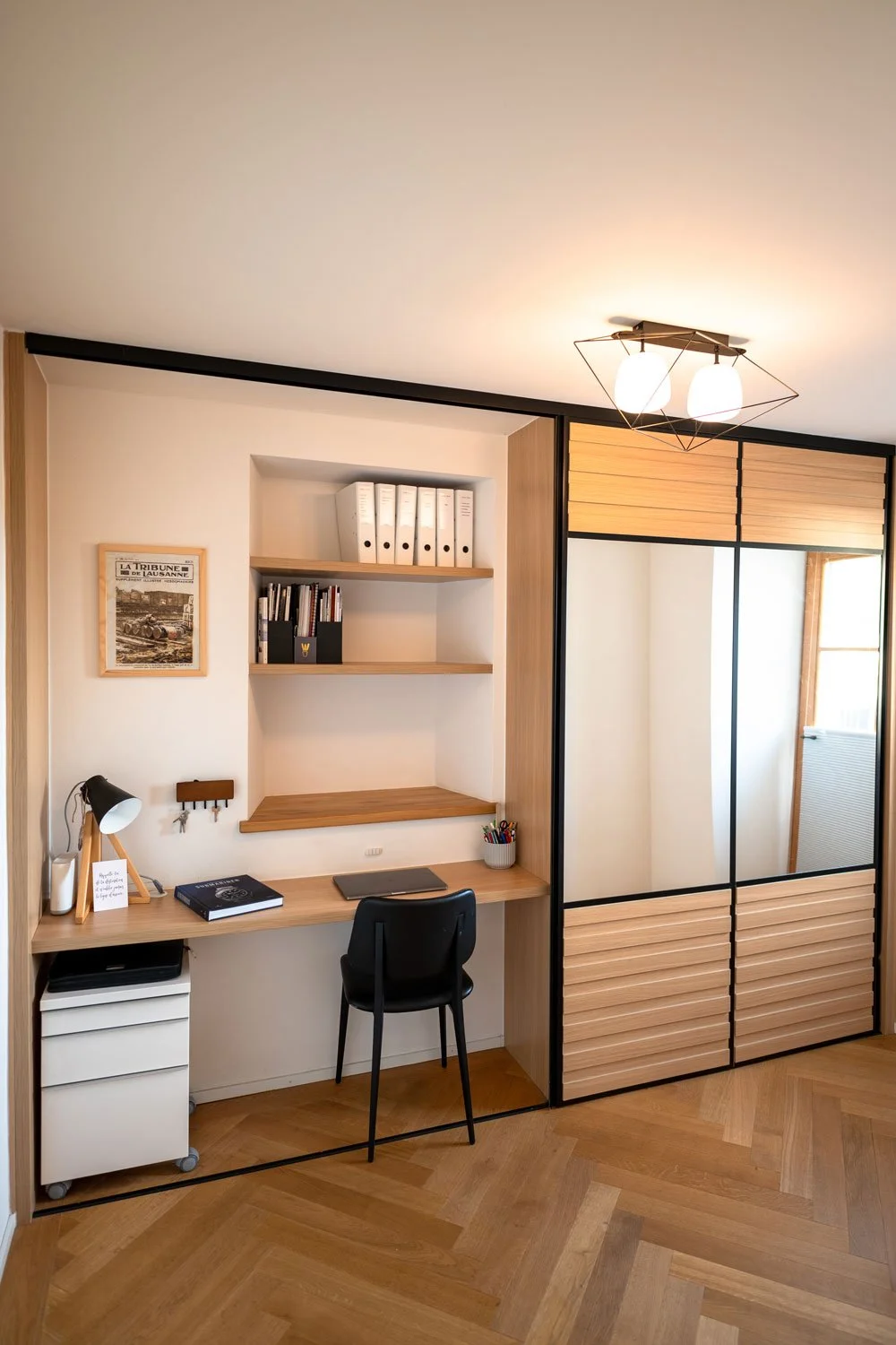 Un coin bureau avec un bureau en bois, une bibliothèque murale avec des classeurs et des livres, un fauteuil noir, une lampe de bureau, un pegboard avec des clés, un poster encadré, un placard avec portes coulissantes en bois et miroir, et un éclairage mural à deux lampes. Sol en parquet bois.