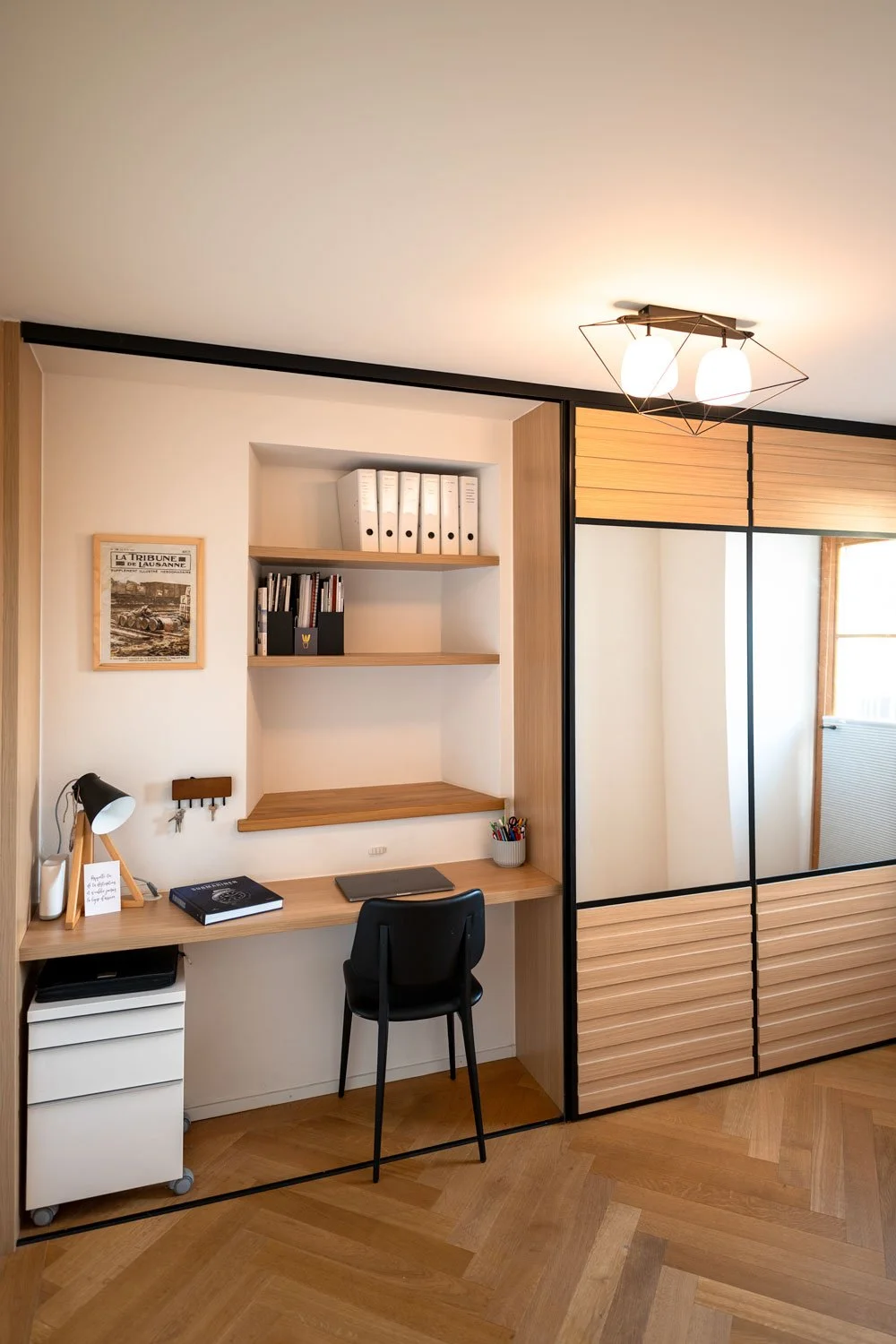 Espace de travail avec bureau en bois, étagères, bibliothèque avec classeurs, lampe de bureau et fauteuil noir, séparé d'une zone avec grand miroir, lumière suspendue et parquet en bois.