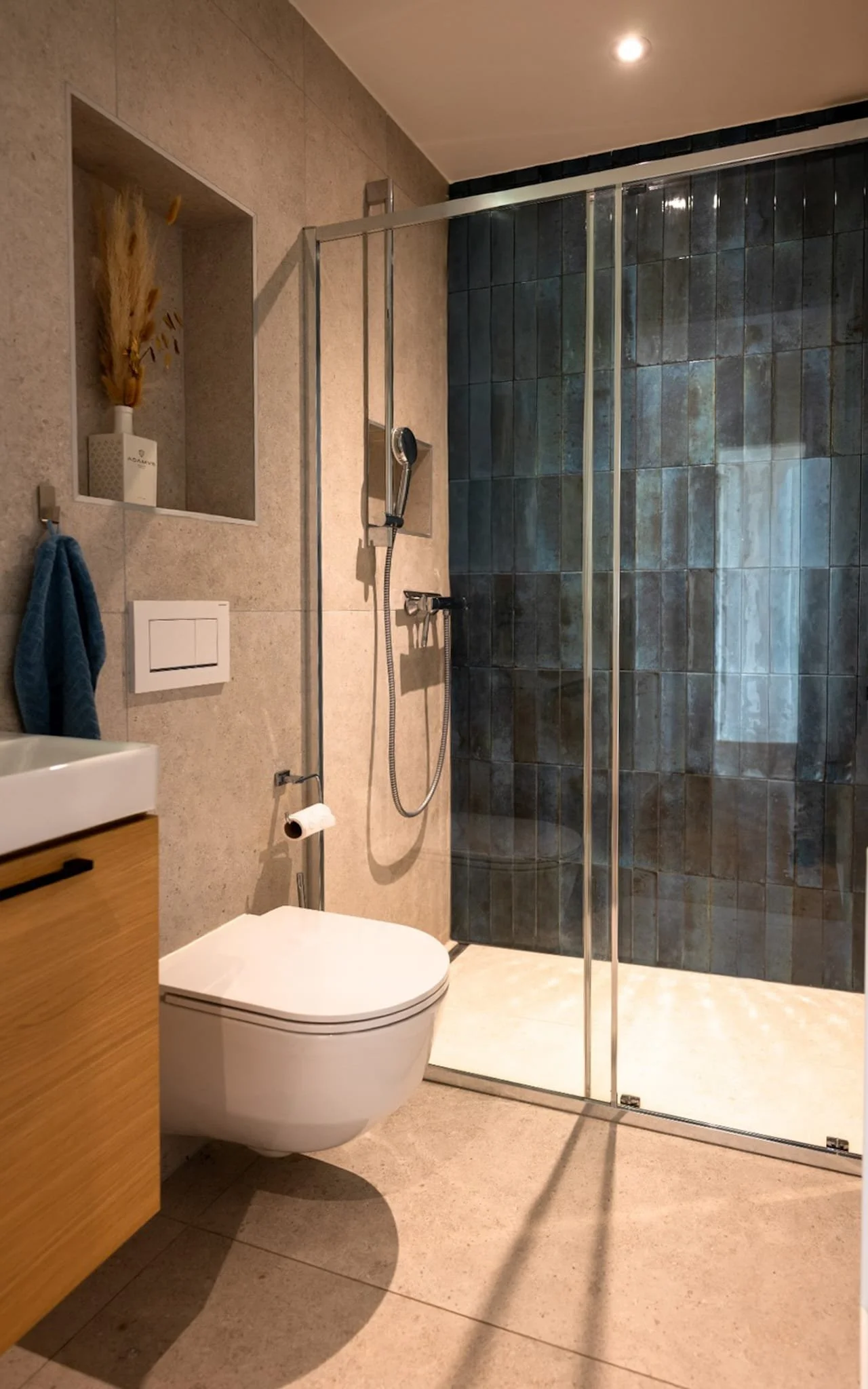 Salle de bain moderne avec WC suspendu, cabine de douche en verre avec carrelage foncé sur le mur, vasque en bois et niches pour rangement.