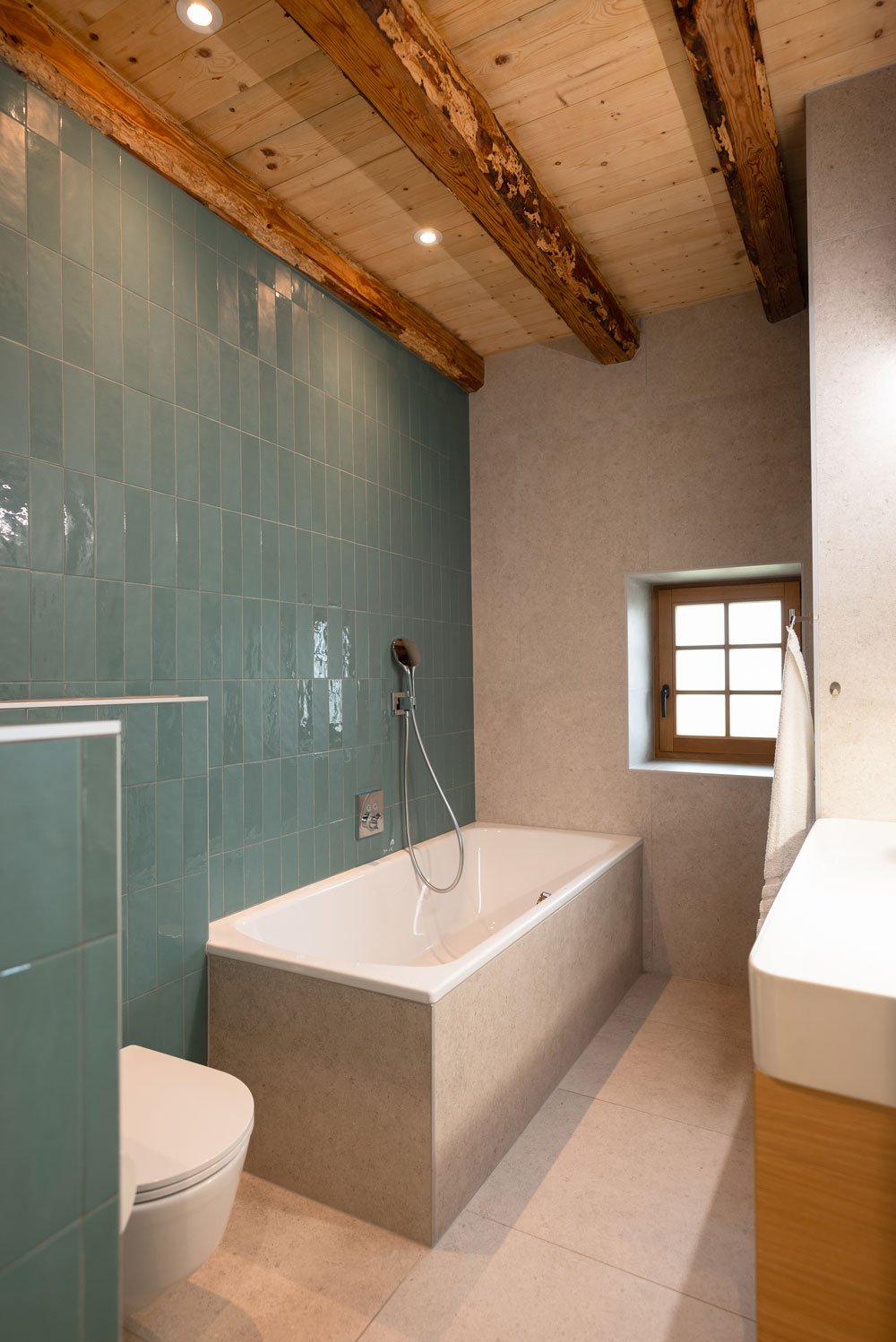 Salle de bain avec baignoire, toilette, lavabo, mur en carreaux verts, plafond en bois avec poutres apparentes, fenêtre en bois, éclairage encastré.