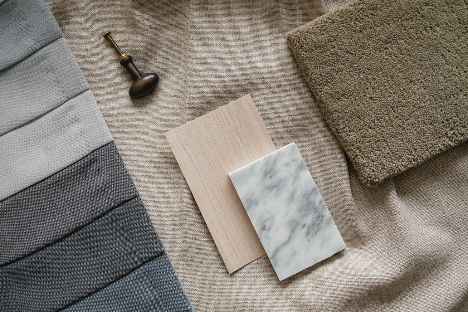 Échantillons de matériaux et textures : tissus en dégradé de gris, échantillon en bois clair, plaque de marbre blanc avec veines grises, tapis beige, poignée de porte en métal ancien, sur une surface en lin beige.