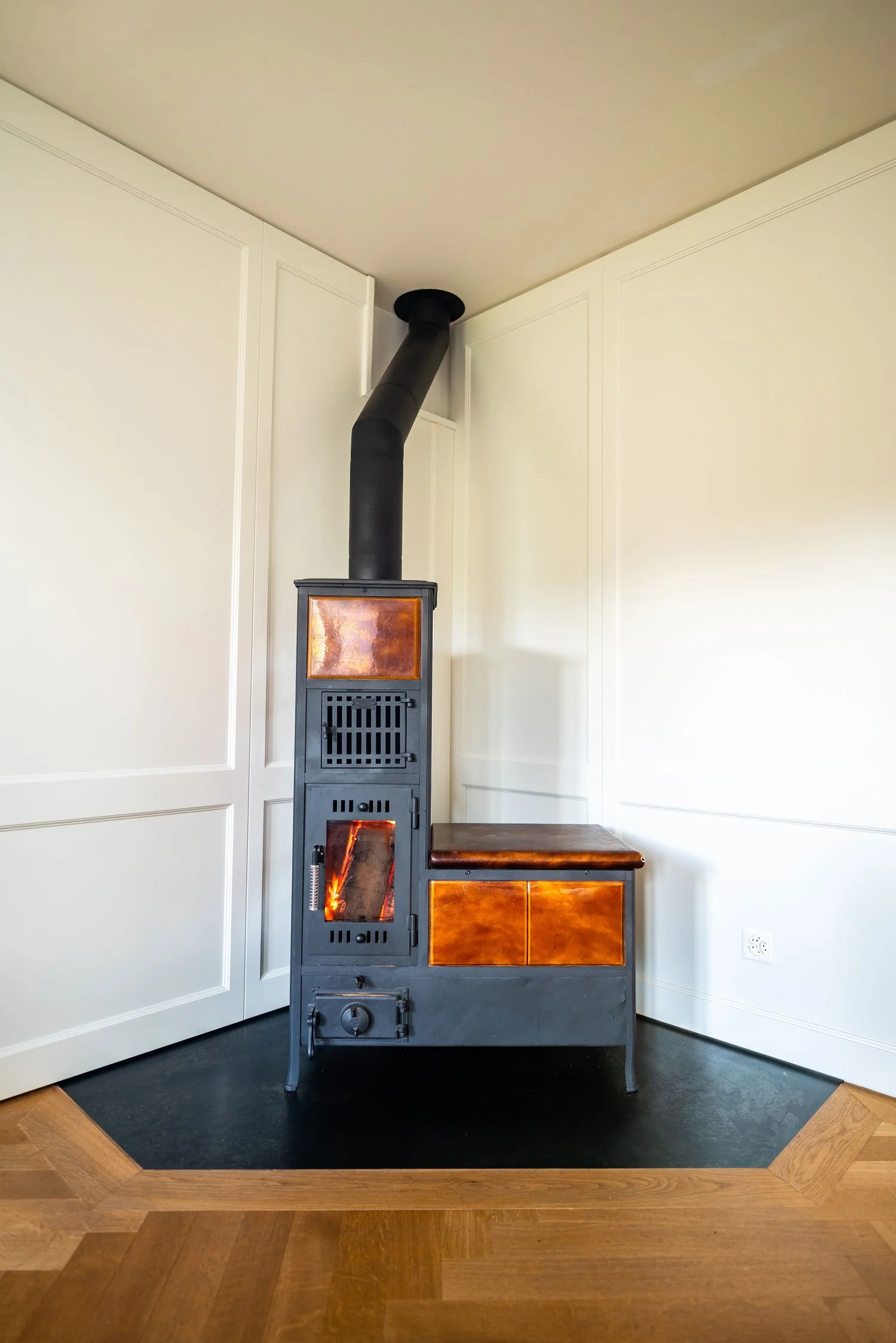 Poêle à bois en métal noir avec portes en verre, placé dans le coin d'une pièce avec murs blanc cassé et sol en parquet, connecté à un conduit d'évacuation noir en haut.
