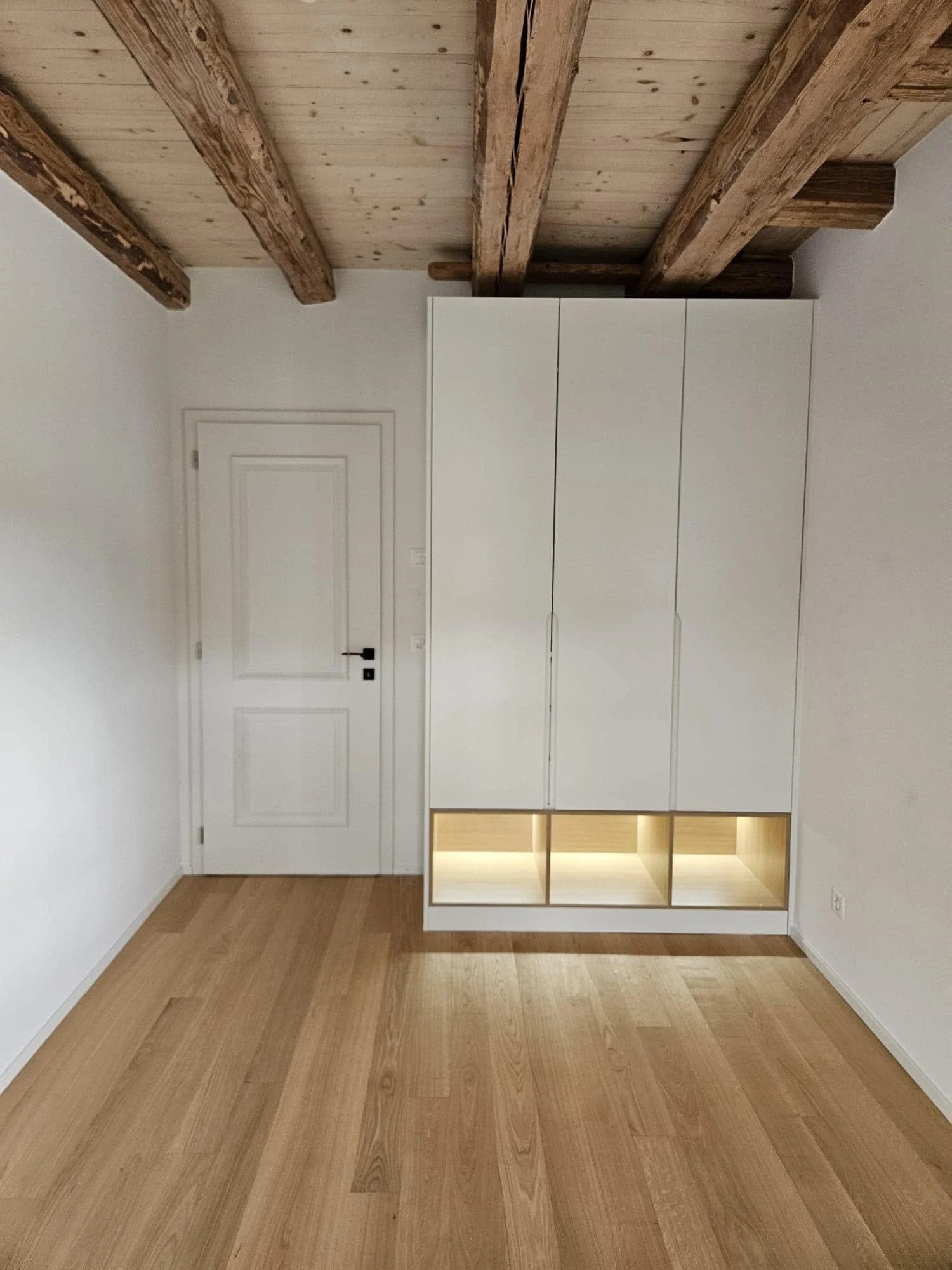 Pièce vide avec parquet en bois clair, mur blanc, porte blanche, armoire blanche avec éclairage intégré et espaces de rangement ouverts en bas, plafond avec poutres en bois apparentes.