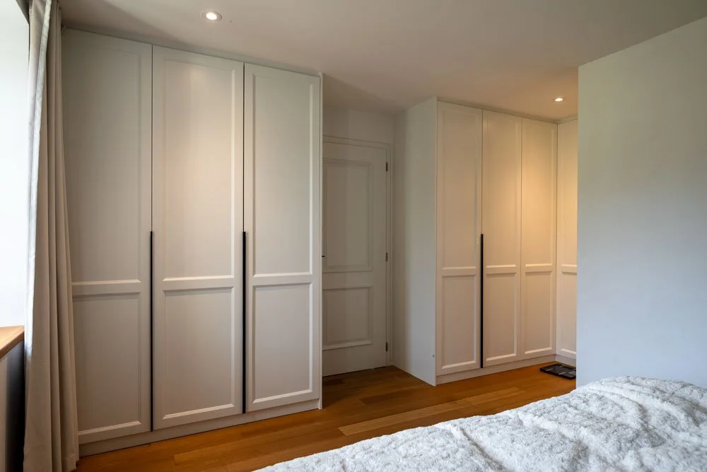 Chambre avec plusieurs armoires blanches, porte en arrière-plan, sol en bois, et lit en premier plan.