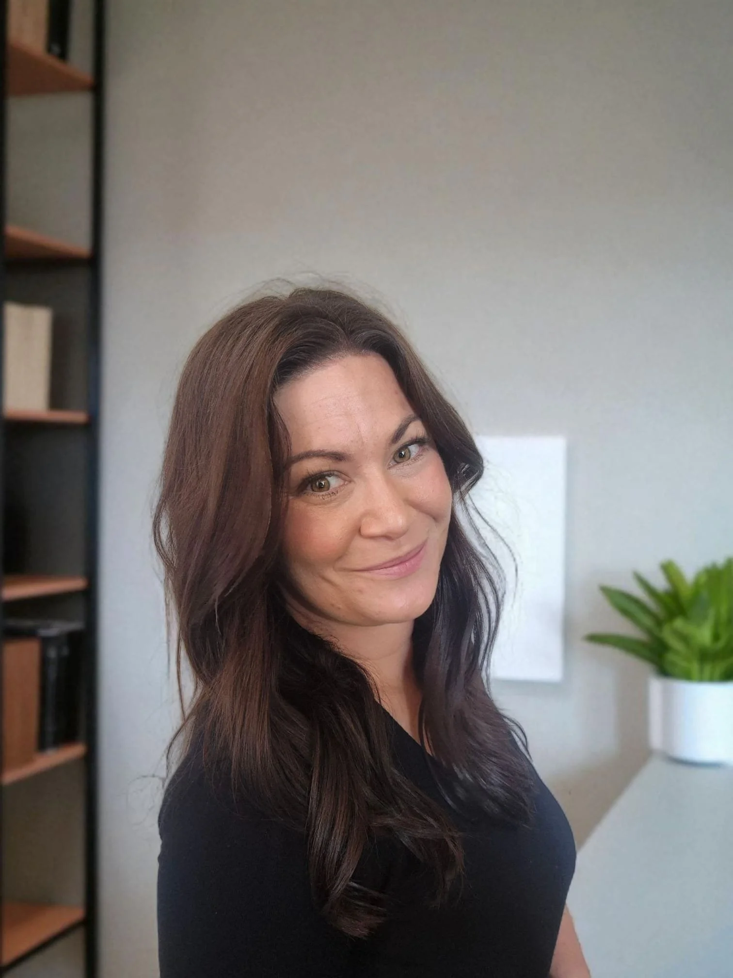 Une femme aux cheveux longs et bruns sourit dans un intérieur domestique, avec une étagère en bois et un pot de plantes vertes en arrière-plan.