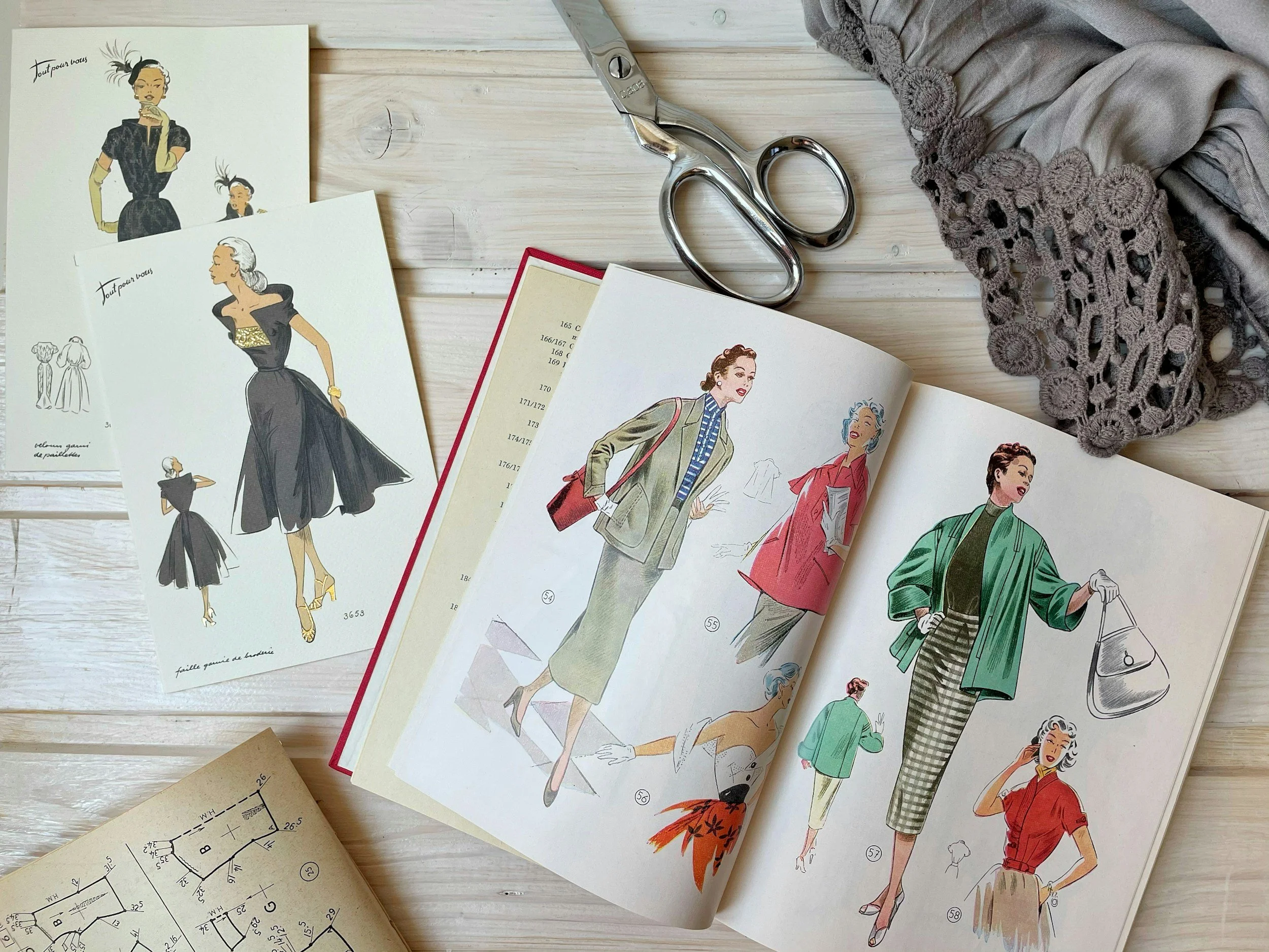 Fashion sketches and illustrations of women in stylish clothing, along with sewing patterns, scissors, and a gray crocheted scarf on a white wooden surface.