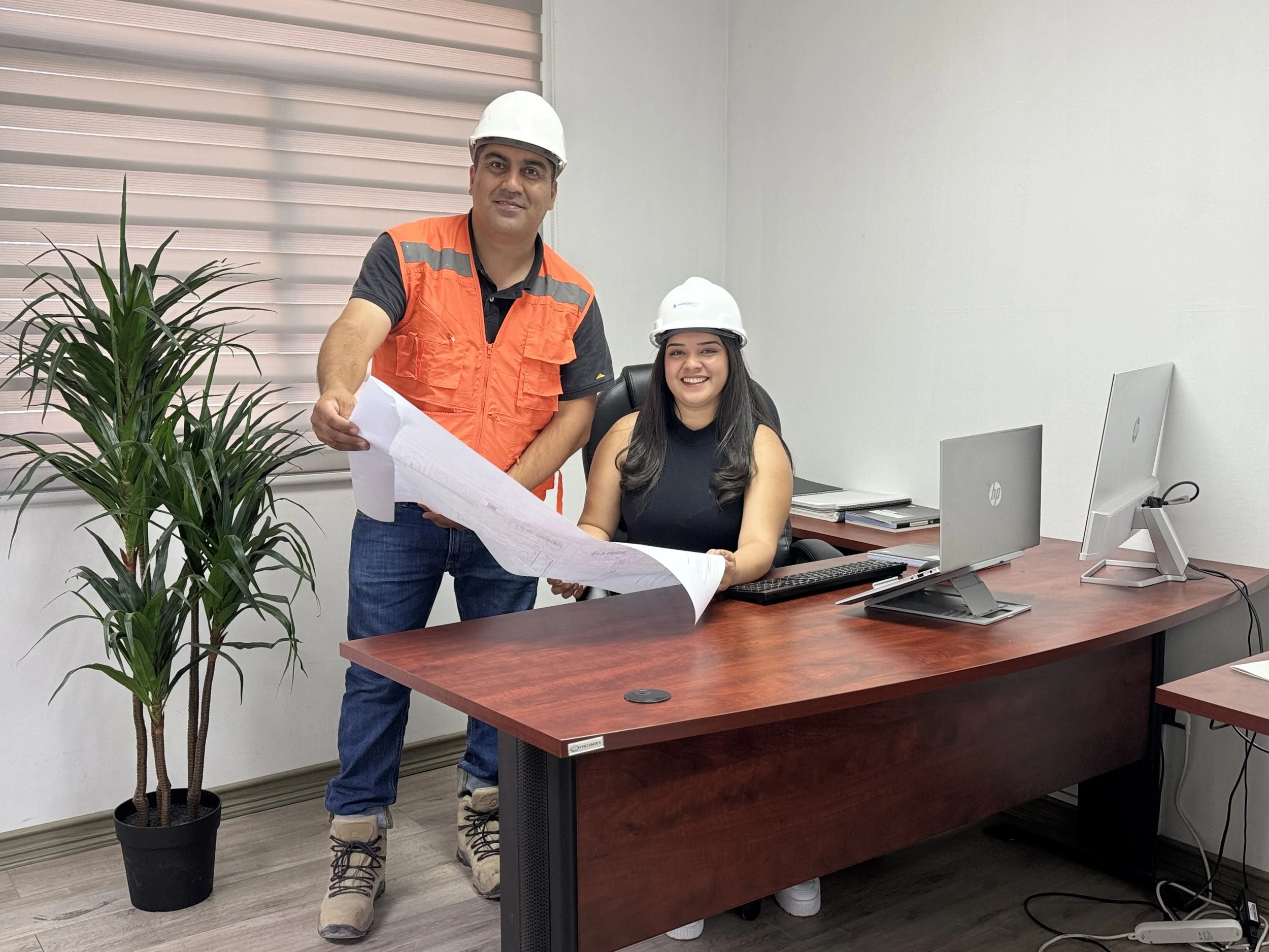 Hombre y mujer en oficina usando cascos de seguridad, la mujer trabaja en una computadora y el hombre sostiene planos, hay una planta y varias computadoras en el escritorio.