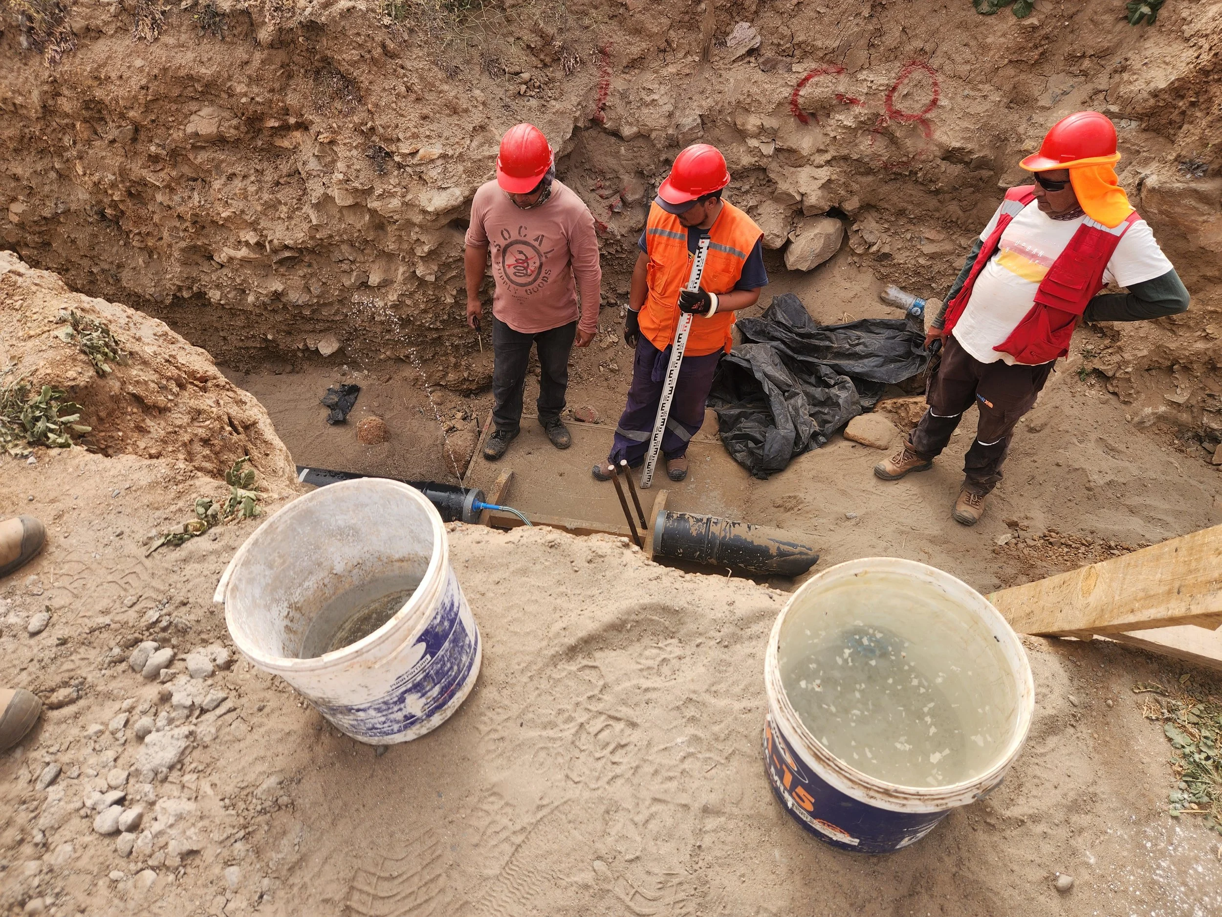 Tres trabajadores en casco y ropa de protección en una excavación, inspeccionando un tubo debajo de la tierra.