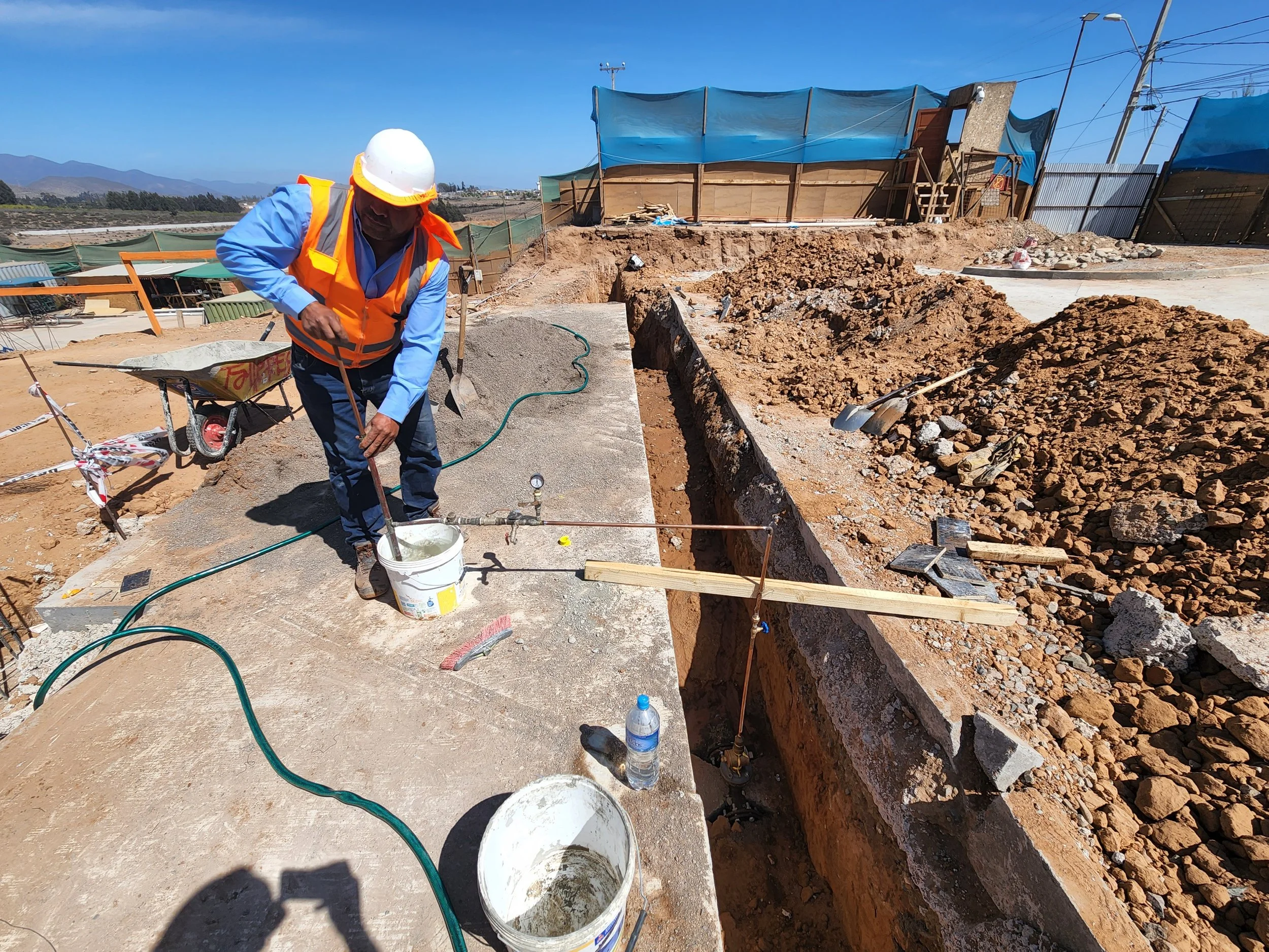 Obrero con casco de seguridad y chaleco naranja trabajando en una obra de construcción en un área al aire libre, con tierra, rocas y herramientas a su alrededor.