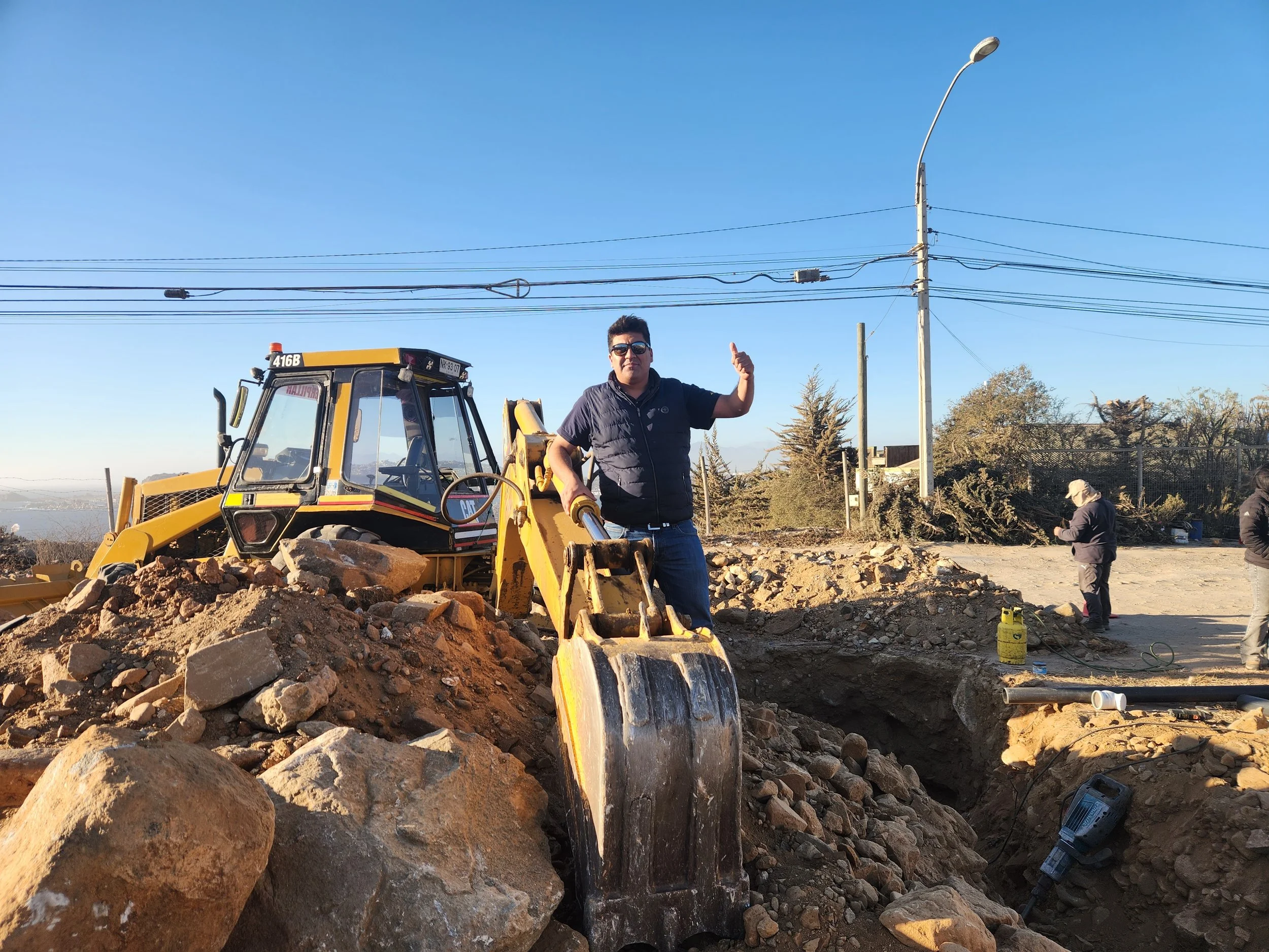 Hombre con gafas de sol junto a una excavadora en un sitio de construcción, fondo cielo azul y algunos árboles y trabajos en proceso.
