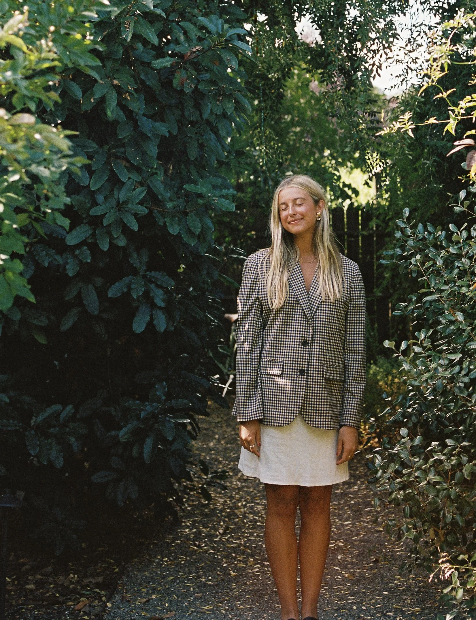 A young woman with long blonde hair standing on a narrow garden path surrounded by lush green bushes. She is wearing a checkered blazer and a white skirt, with her eyes closed and a gentle smile on her face.