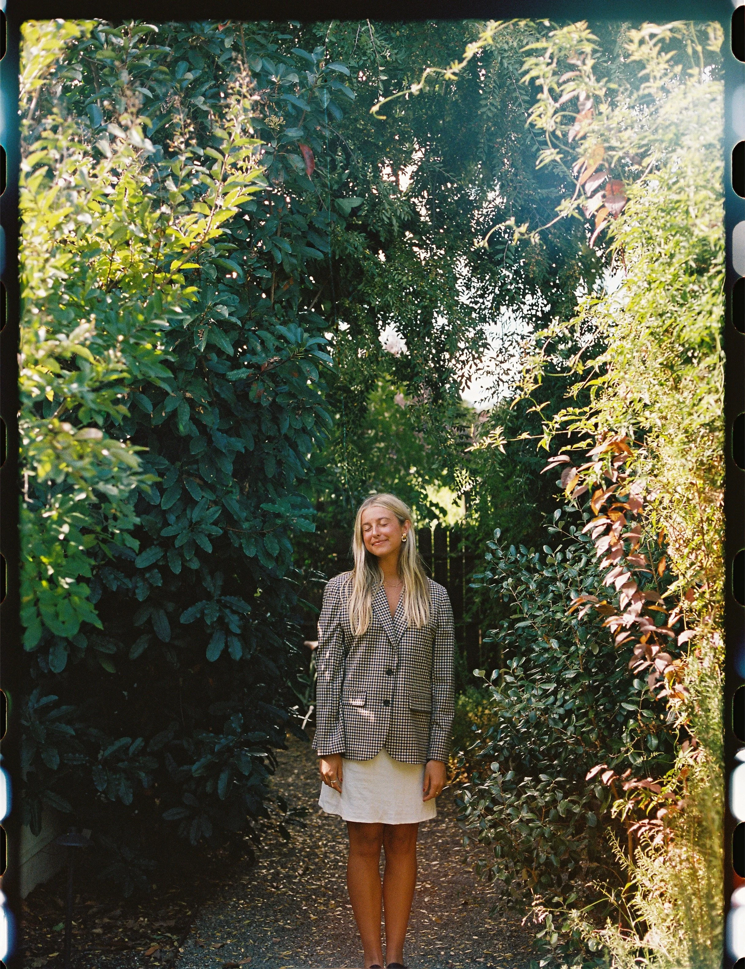 A woman with long blonde hair wearing a checked blazer and a white skirt standing on a narrow garden path surrounded by lush greenery.
