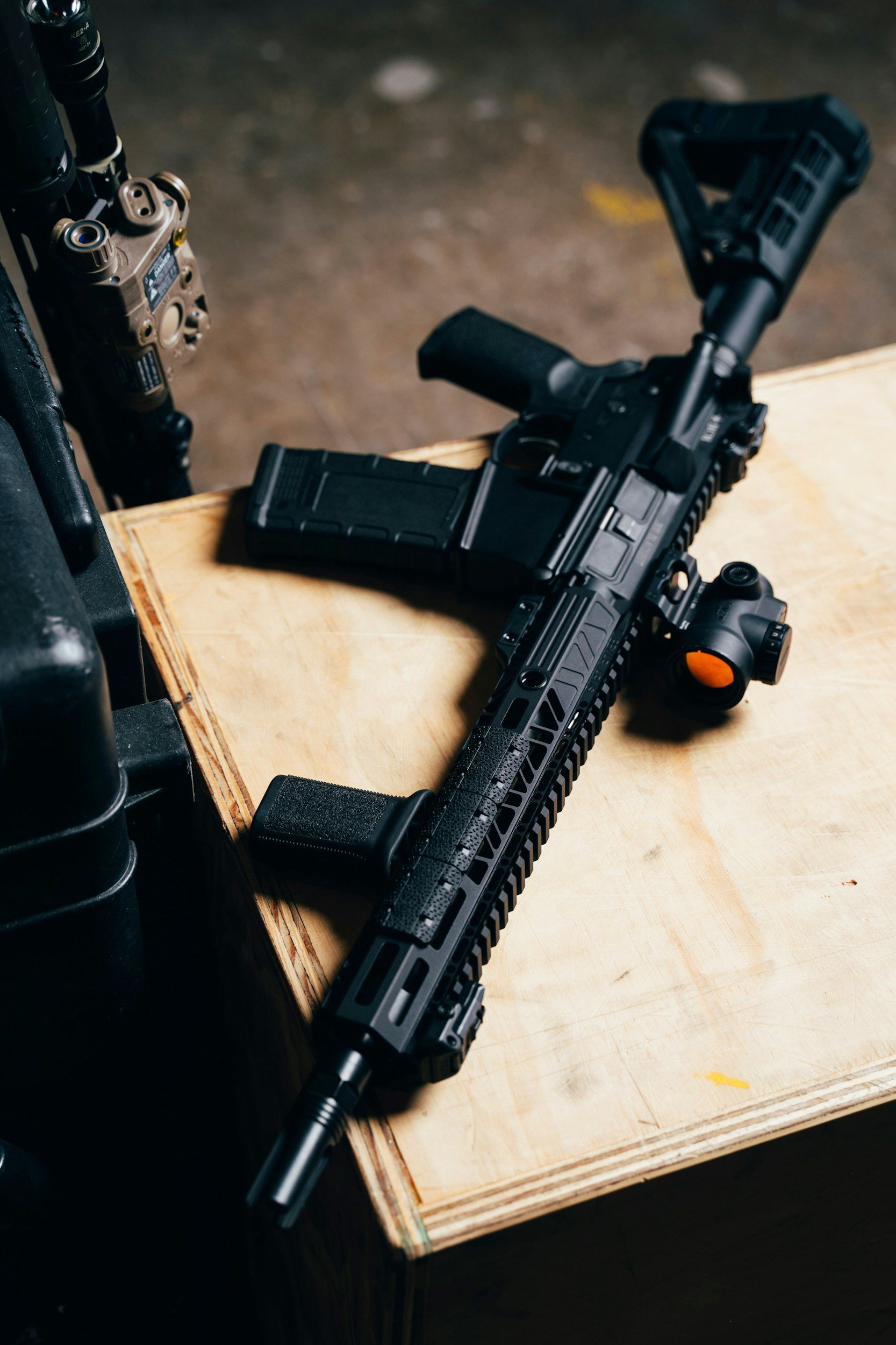 A black tactical rifle with a scope and a mounted magazine lies on a wooden surface in a dimly lit setting.